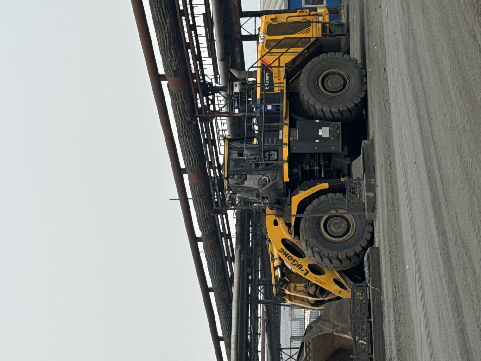 A large, yellow front-end loader, branded with "LU GONG" and "5.973" on its side, is parked on a gravel surface beneath an elevated structure with multiple large, rust-colored pipes. The loader is positioned centrally in the frame, facing right. Its large tires and bucket are visible. The environment appears industrial, with muted colors and an overcast sky. The background shows a grey, possibly industrial, building with a blue section. The scene is static with no people or animals present. The location is identified as Norilsk, Russia.