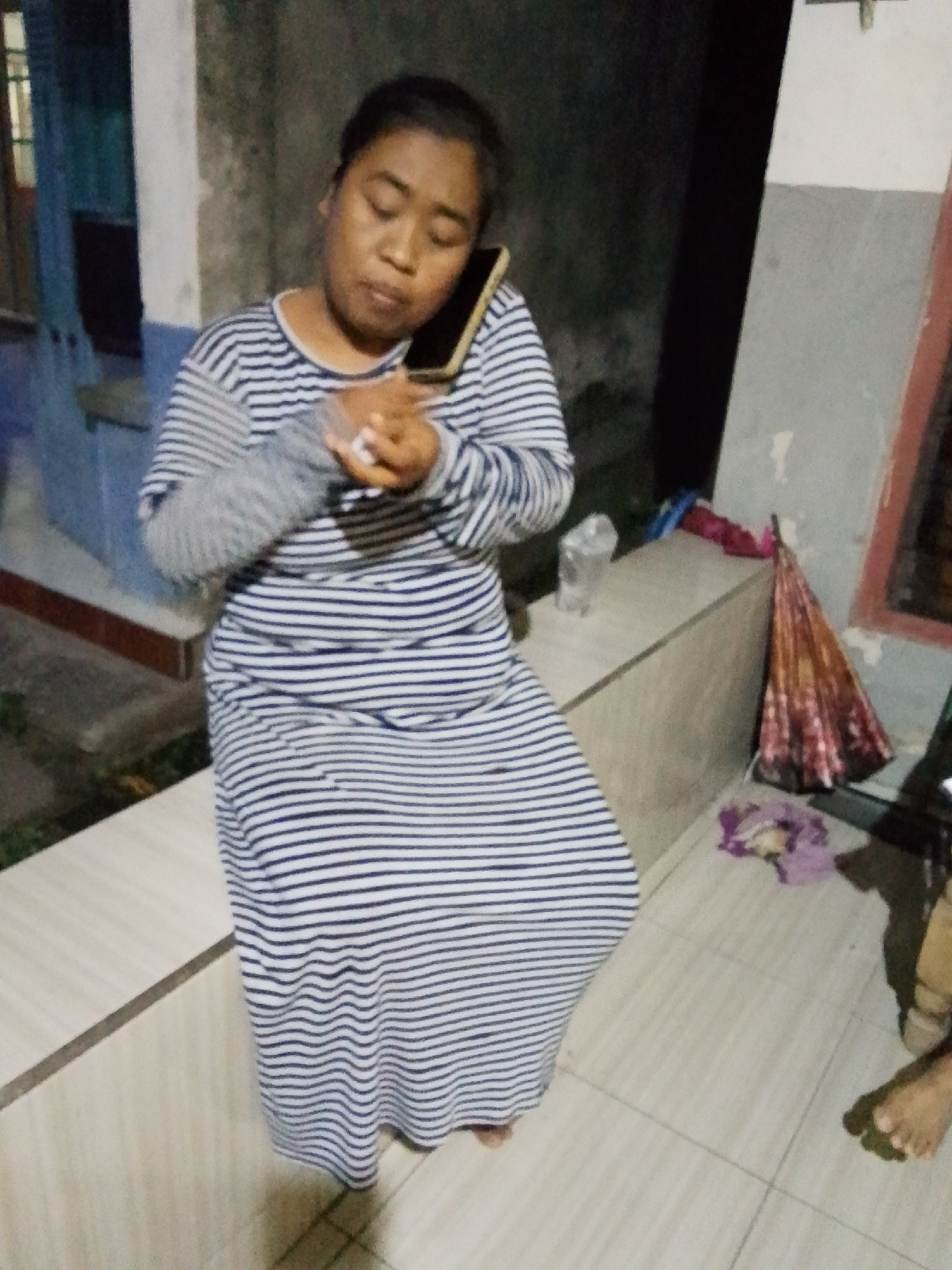 In Bulurejo, Indonesia, a person is sitting on a ledge and talking on a mobile phone. They are wearing a long-sleeved, horizontally striped dress. In their right hand, they are holding a small white object. To the right of the person, a folded umbrella with red and orange patterns rests on a surface. The environment appears to be outdoors, with tiled flooring and textured walls.