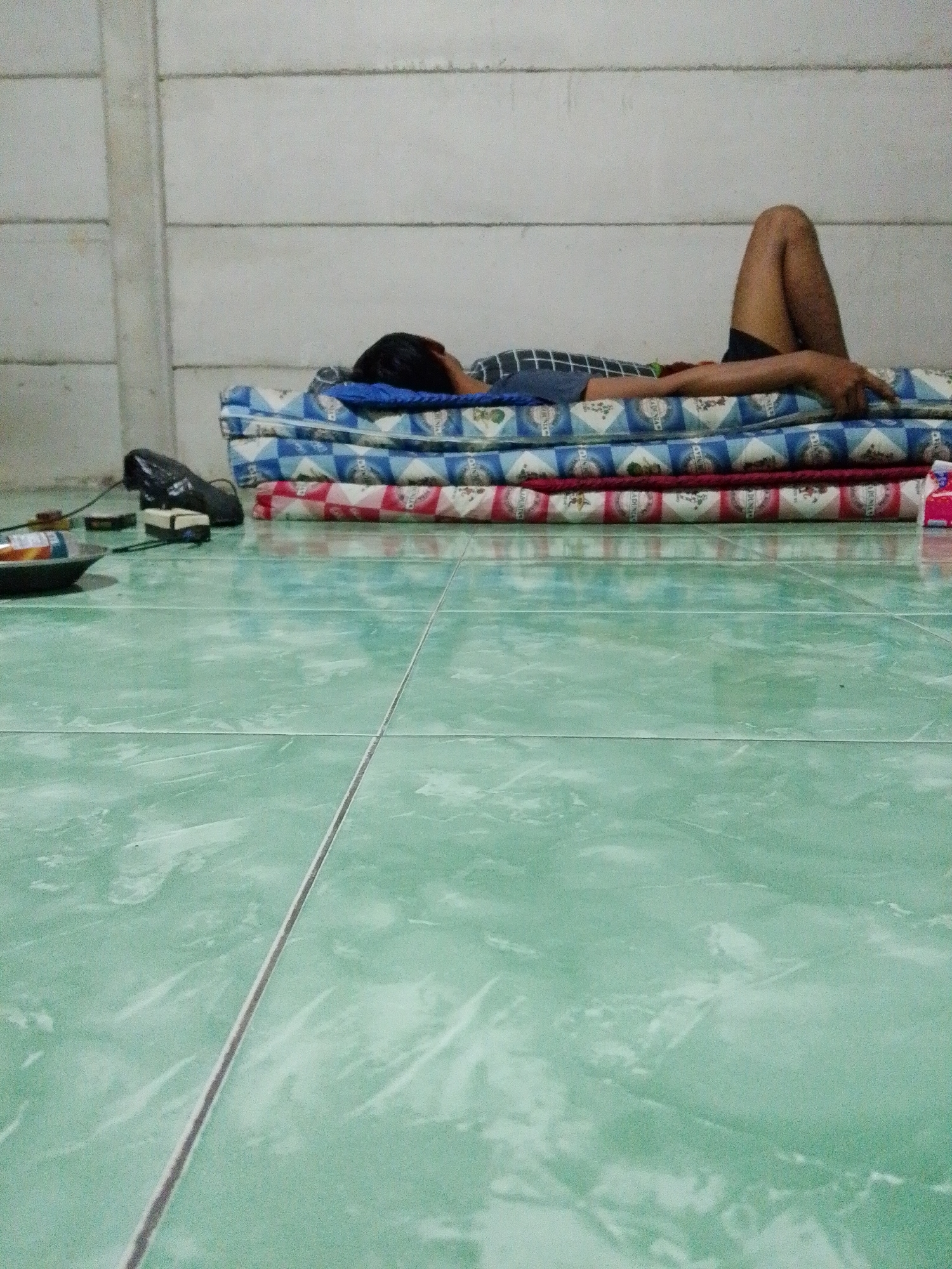 A person is lying on a stack of mattresses on a tiled floor in Bukit Ketok, Indonesia. The person is lying on their back with one leg bent at the knee, the other leg extended. The floor is green tile. A bag and some other objects are on the floor near the mattresses. The wall is white.