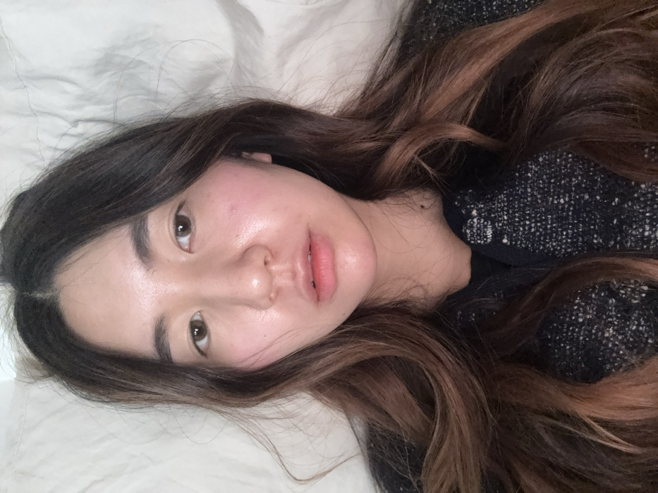A close-up portrait shows an East Asian female with long, wavy dark brown hair featuring reddish-brown ombré highlights. Her brown eyes are open, and she gazes directly forward with a neutral expression, her lips slightly parted. Her skin shows some natural redness on the cheeks and a subtle sheen on the forehead. She is dressed in a dark, textured outerwear garment, possibly a tweed-knit jacket or cardigan, over a visible black undergarment. The immediate background consists of a crumpled, plain white or light-colored fabric, indicative of bedding. No specific environmental details are present to determine a precise location within the city of Paris, France, or any other geographical area.