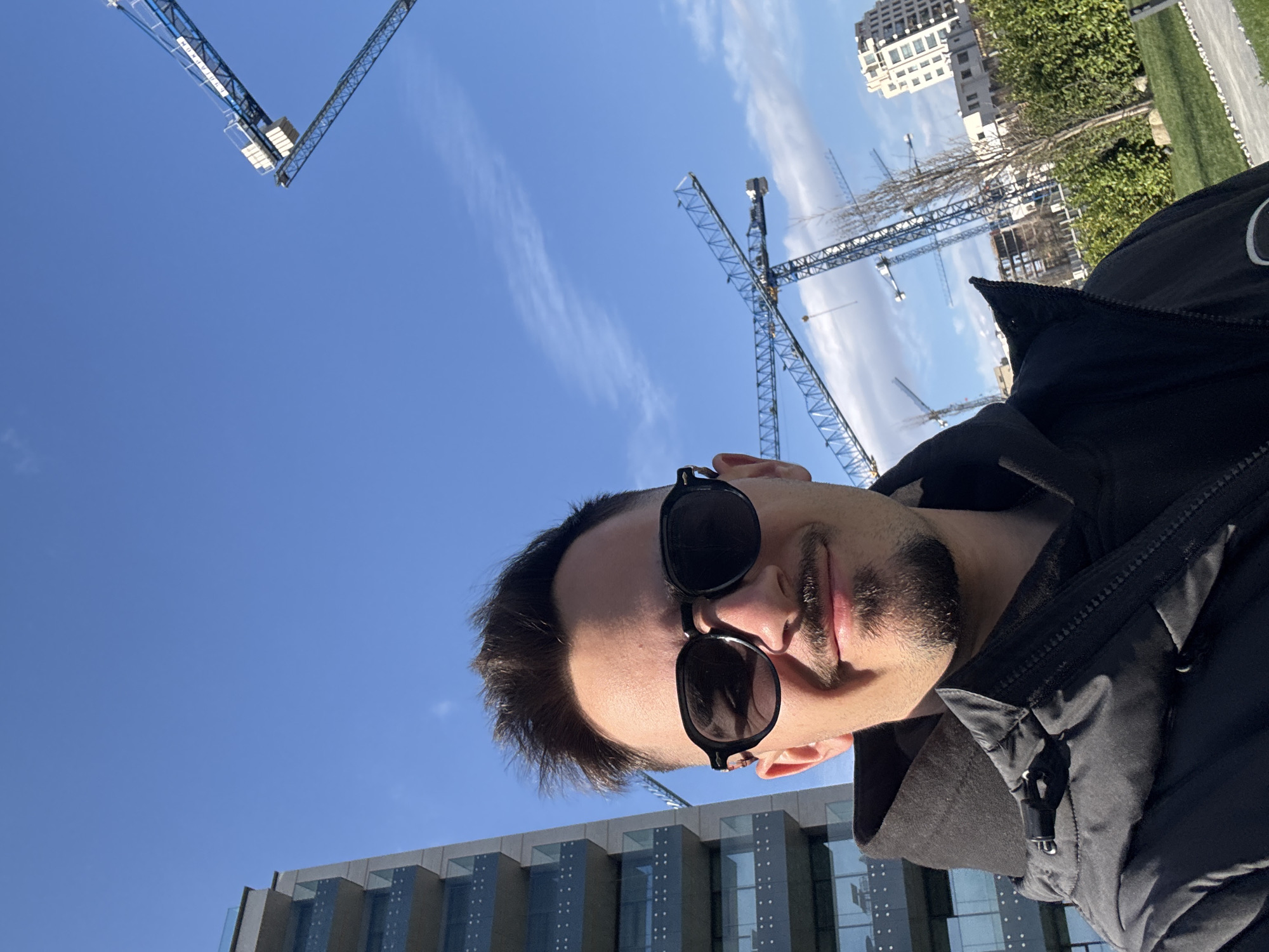 A selfie of a man with sunglasses on a sunny day in Sofia, Bulgaria. The background shows multiple construction cranes and a few buildings. The sky is blue with scattered clouds. The man is wearing a dark jacket.