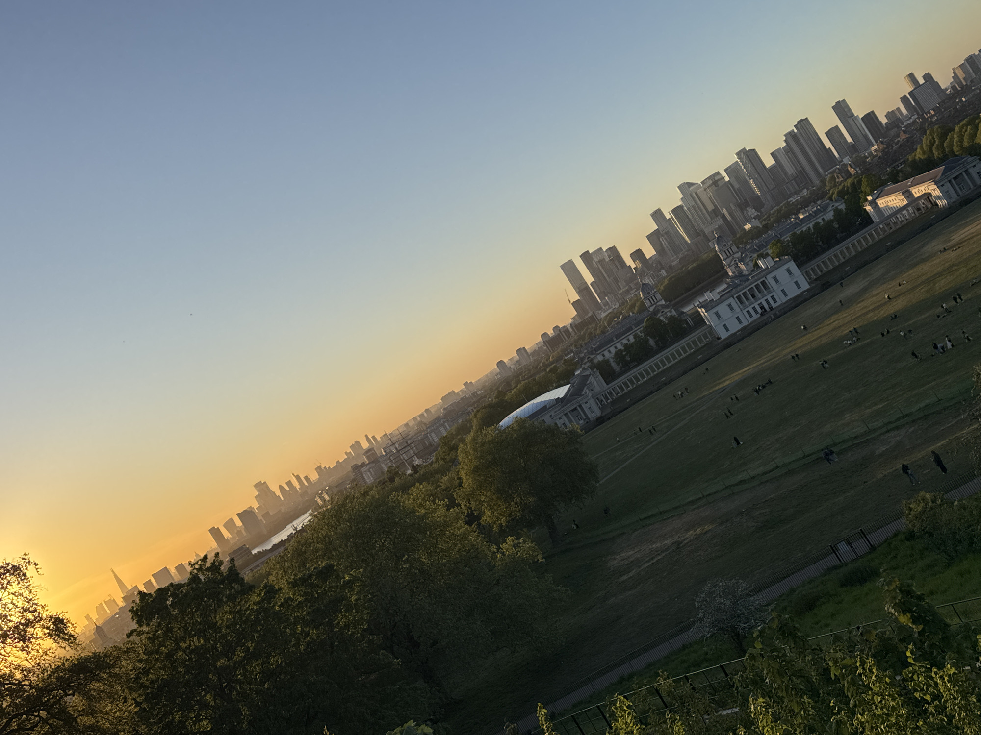 This panoramic image, captured at an oblique angle, presents a scenic view from an elevated position, likely Greenwich Park, overlooking parts of London during sunset or late afternoon.
In the foreground, lush green trees frame the bottom and left side of the shot, silhouetted against the bright sky on the left. A wide expanse of green parkland dominates the middle ground, where numerous small figures of people are scattered, engaging in leisure activities such as sitting, walking, and relaxing. A distinct, large white classical building with a dome and colonnades, identifiable as part of the Old Royal Naval College, stands prominently within the park area.
Beyond the park and the historic buildings, a modern urban landscape unfolds. To the right, a dense cluster of tall skyscrapers forms the distinct skyline of Canary Wharf. Further to the left, in the hazy distance, another iconic city skyline is visible, including the recognizable silhouette of The Shard. A body of water, likely the River Thames, separates these two urban clusters.
The sky transitions from a soft blue at the top to a warm, vibrant orange and yellow near the horizon, indicating the time of day is approaching sunset. The sun, partially obscured by trees on the far left, casts a warm glow, silhouetting the distant buildings and creating a serene atmosphere. The weather appears clear and calm, enhancing the picturesque quality of the scene. No discernible text is visible in the image.