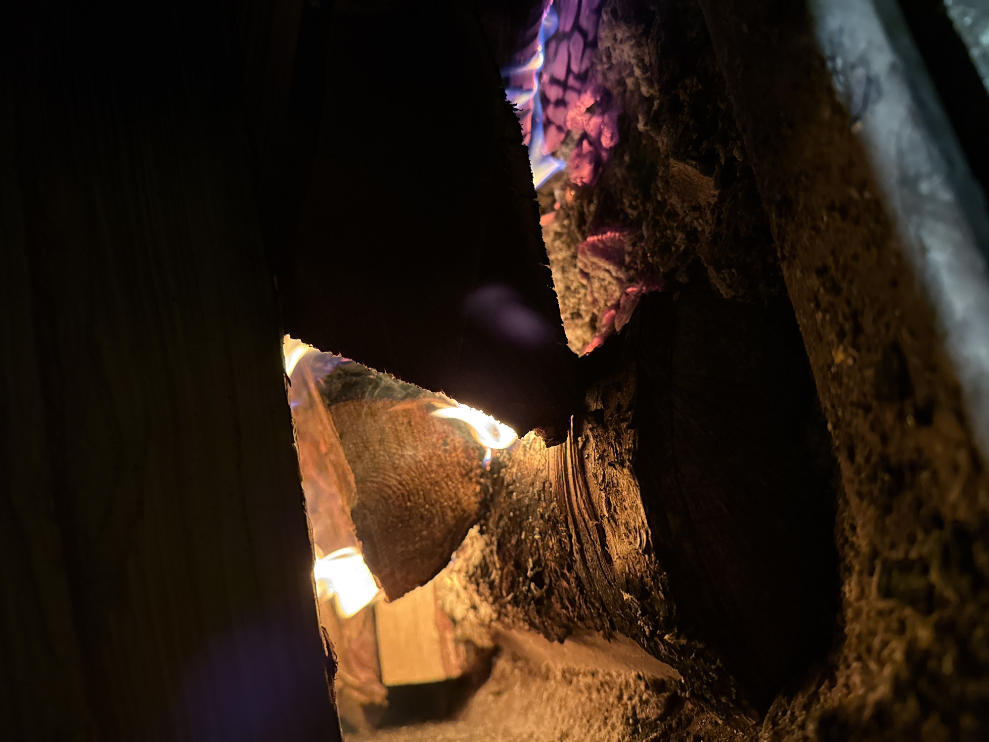 A close-up image shows burning logs in a fireplace or wood-burning stove. The logs are arranged in a way that creates gaps and allows the fire to be seen. The flames are visible through these gaps, with hues of orange, yellow, and blue. The logs themselves appear charred and textured. The surrounding environment is dark, suggesting the image was taken indoors with minimal lighting. There is no verifiable information to confirm this image was taken in Tyulyuk, Russia.