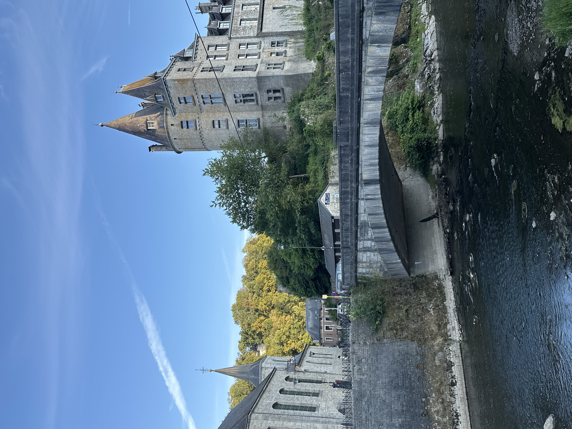 This is a photograph of the town of Durbuy, Belgium. The foreground features a dark, rippling river with visible rocks and some green algae. A stone bridge with a concrete underside arches over the river, with a grey stone wall on the left bank. Behind the bridge, a prominent stone building with conical towers, resembling a castle, is situated on a grassy incline. To the left of the castle is a stone church with a tall, pointed spire. In the background, a dense forest with autumn foliage covers a hillside. The sky is a clear blue with a faint contrail visible. Several vehicles are parked along a road in the middle ground, and some small buildings are nestled between the church and the castle.