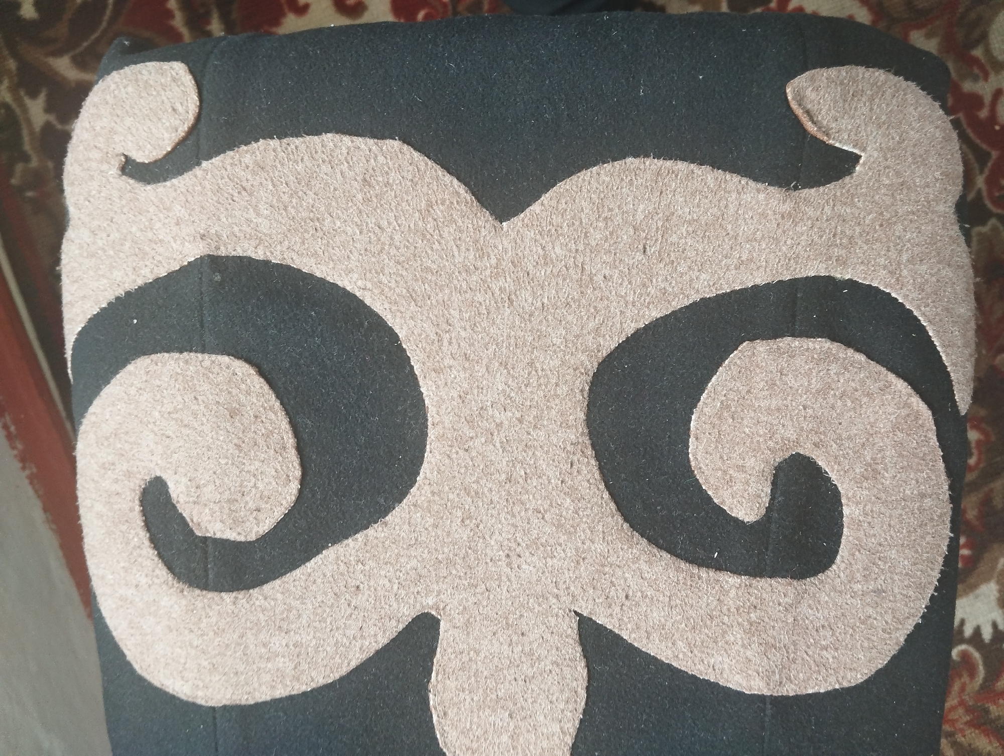 The image shows a close-up view of a black fabric surface with a beige, felt-like applique design. The design features symmetrical, swirling patterns that create a stylized face or mask-like appearance. The background appears to be a patterned rug or carpet with red and brown tones. The scene is indoors, and the lighting is even. There are no people or specific landmarks visible to indicate a location in Naryn City, Kyrgyzstan.