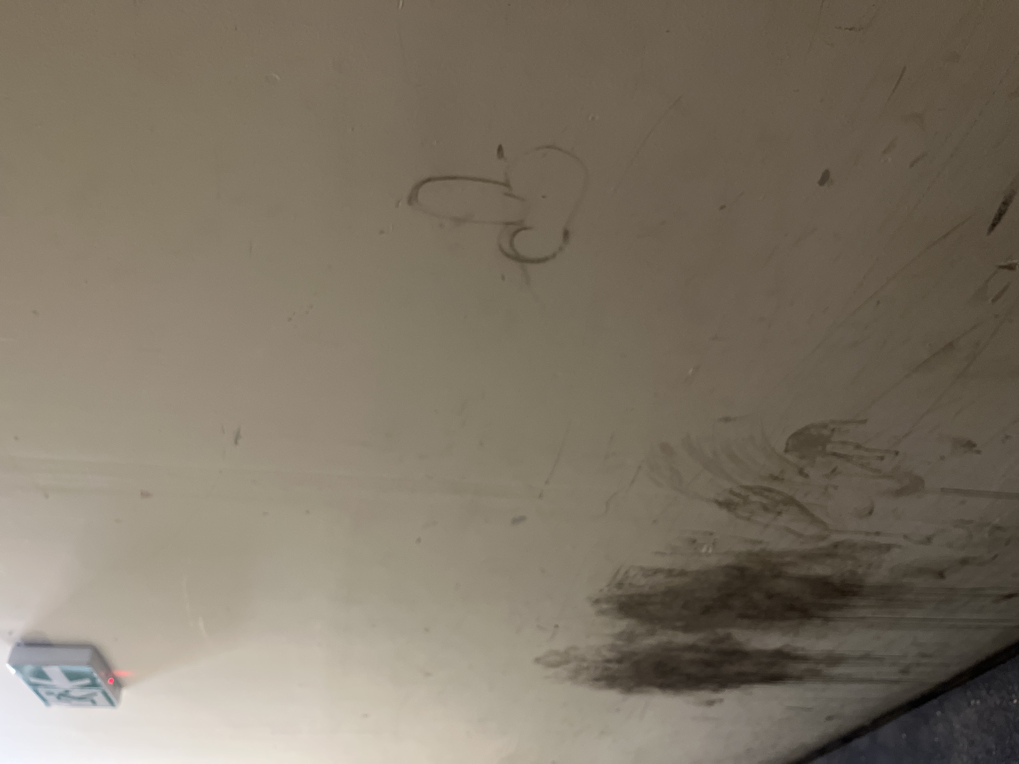 The image shows a light-colored, possibly white or off-white, surface with various markings. In the top center, there's a small, crude drawing. Towards the bottom right, there are darker, smeared markings, possibly dirt or residue. In the bottom left corner, there's a small, rectangular object with a red light emitting from it.
Without additional context, it is impossible to pinpoint the location in Plovdiv, Bulgaria, or provide a more detailed forensic analysis.