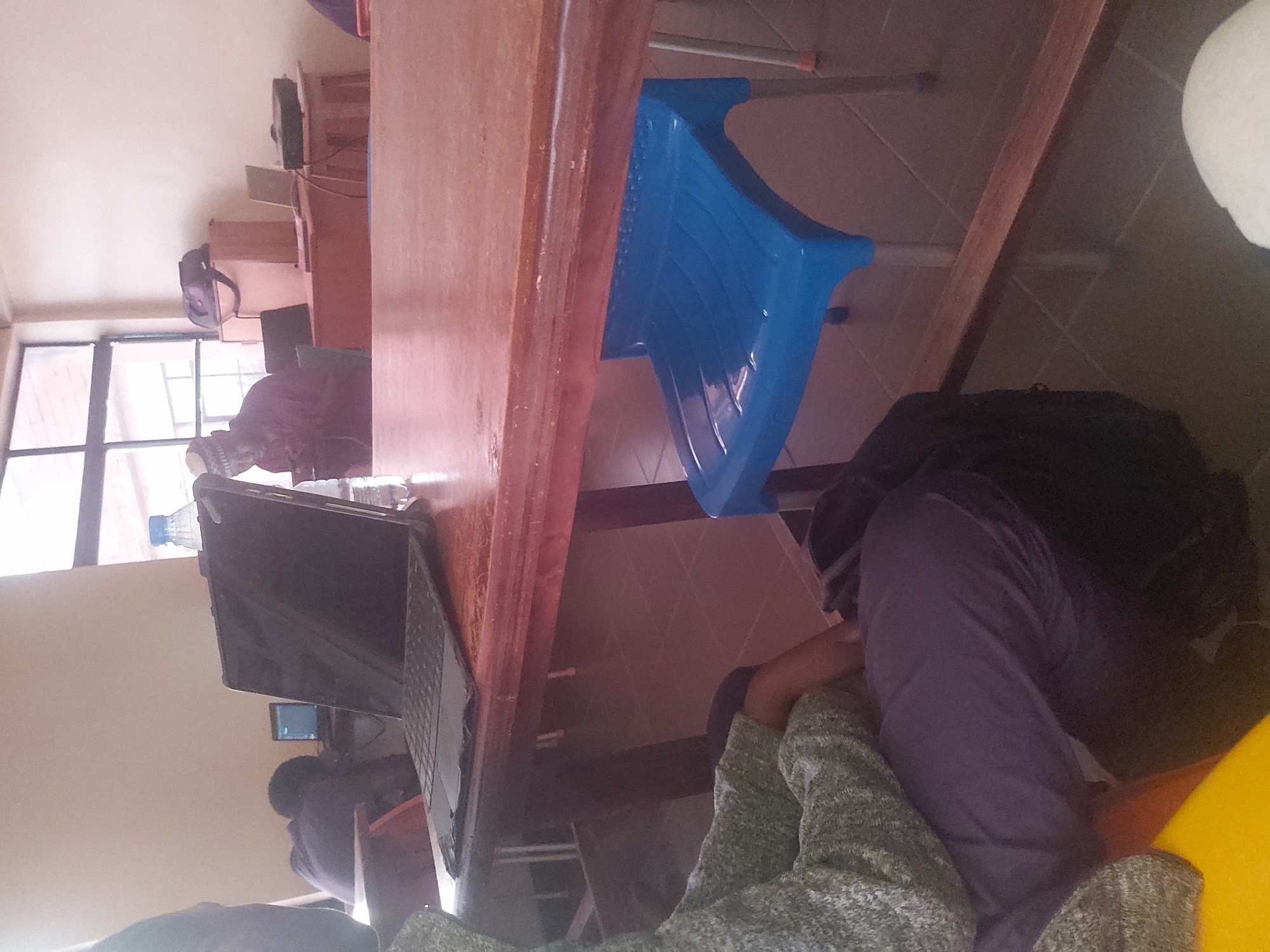 The image depicts an indoor scene, possibly a classroom or study area, with several individuals and objects present. In the foreground, a person is partially visible, wearing a grey sweater and purple trousers. They are seated at a wooden table, on which a black laptop is placed. The laptop appears to be open and in use. To the right of the laptop, a blue plastic chair is positioned under the table. In the background, another person is seated at a desk, facing away from the camera and appears to be using a laptop. Further in the background, more tables and chairs are visible, along with a window with blinds. The overall environment suggests a functional space for learning or work. The presence of a laptop, books, and a projector indicates an educational setting. The tiling on the floor and the lighting suggest an interior location. However, there are no discernible elements that specifically place the scene in Jos, Nigeria.