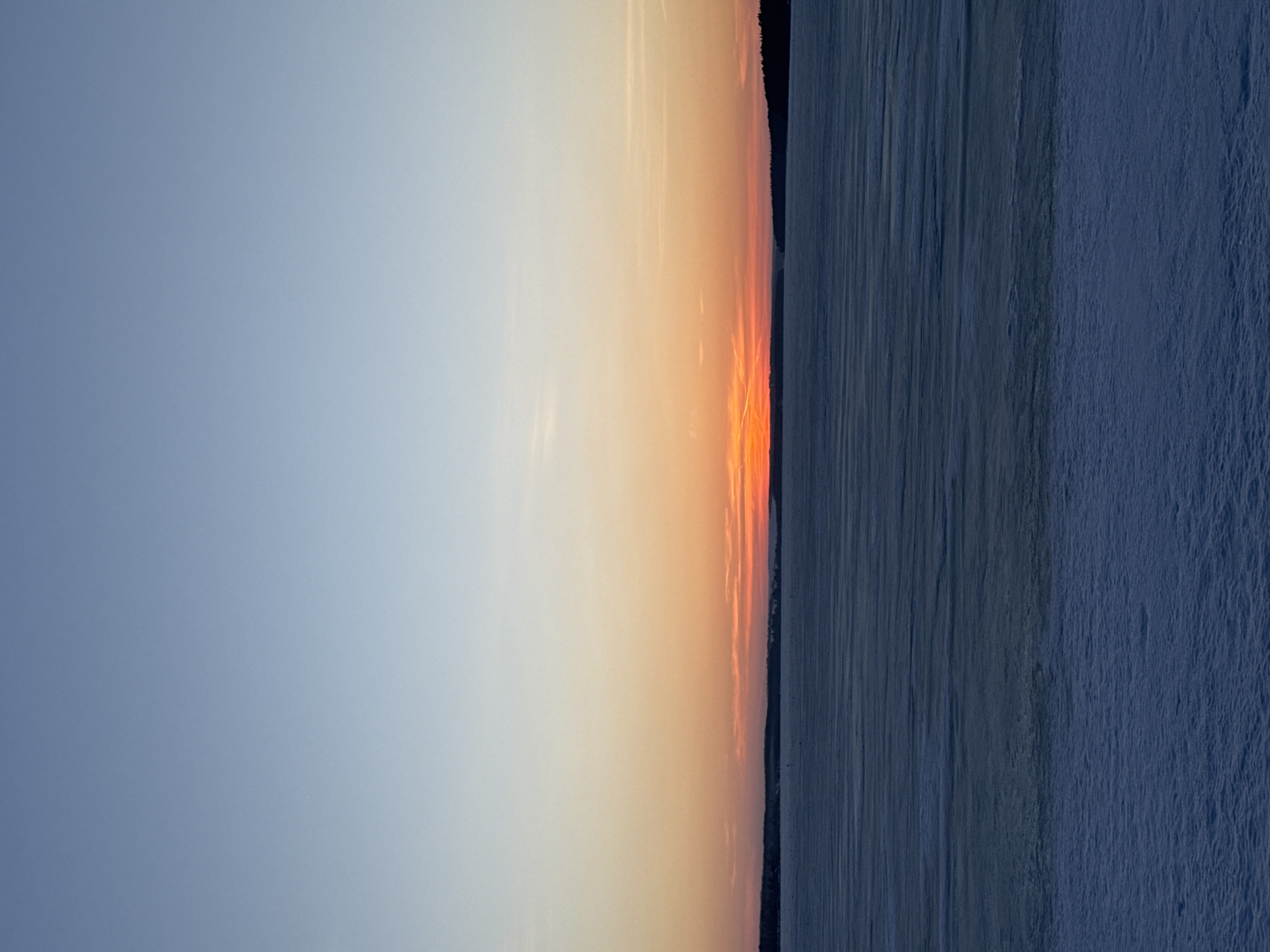 The image captures a serene sunset scene, likely over a body of water, possibly near the city of Votkinsk, Russia, though specific landmarks confirming the exact location are absent. The sky transitions from a pale blue on the left to warm hues of orange and yellow where the sun appears to set on the horizontal center of the image. A faint dark silhouette of land or trees is visible between the sunset and the water. The water or ground in the right half of the image shows a textured, blueish-gray surface, which could be a frozen lake or snow-covered land. No people, specific actions, or interactions are visible. The scene presents a tranquil and possibly cold environment during sunset.