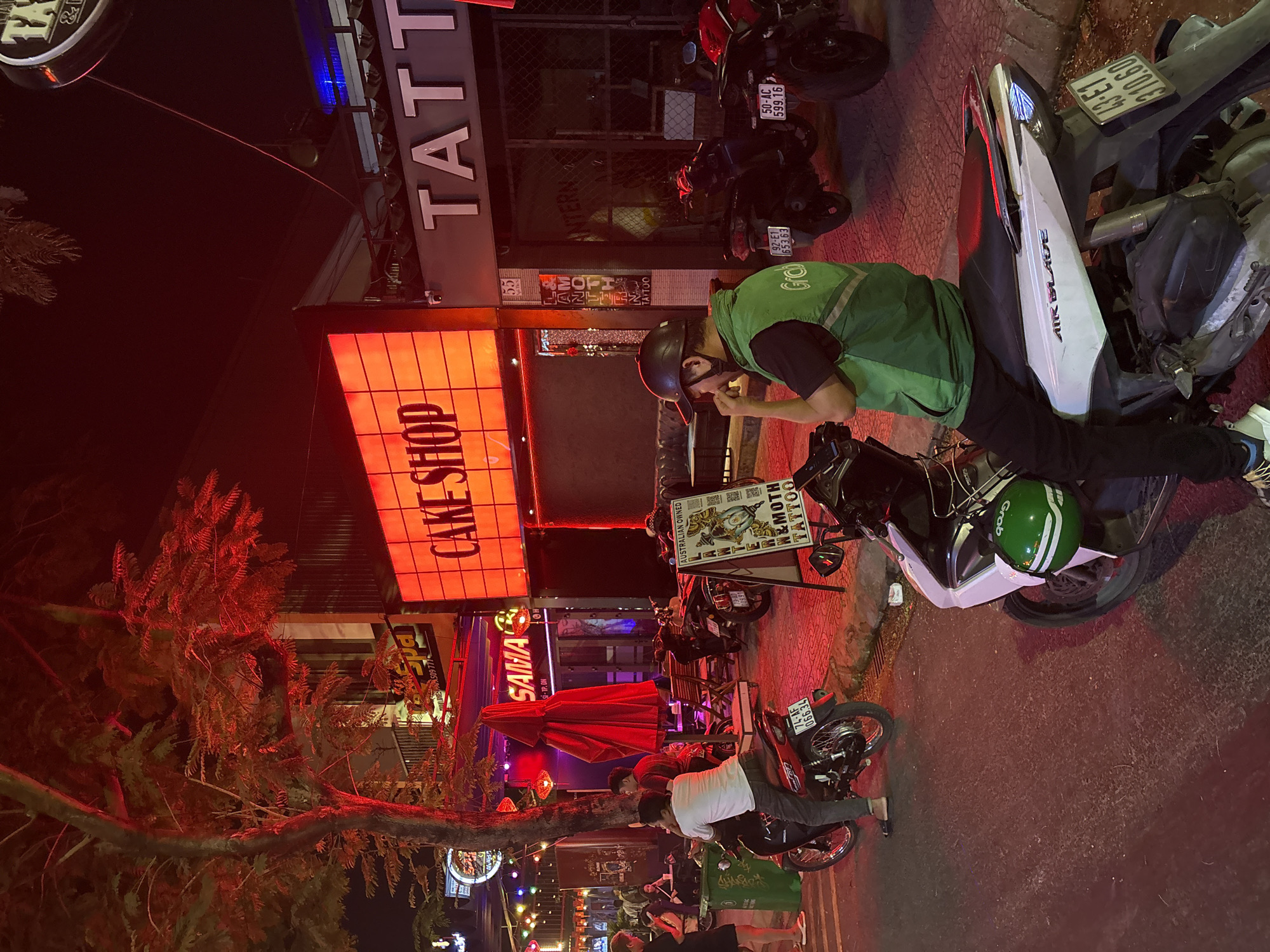 The image depicts a vibrant street scene in Đà Nẵng, Vietnam, at night. A man wearing a green Grab uniform and helmet is standing next to his white motorcycle, possibly eating. The motorcycle's license plate reads "43-E1 310.60". Nearby, another motorcycle has a license plate "74-AF 066.34". The street is lined with various businesses, including a "Cake Shop," a "SAMA" restaurant, and a "N & Moth Tattoo" parlor. The tattoo parlor sign advertises "Australian Owned." Various motorbikes are parked along the street. People are present on the sidewalk. The area is brightly lit with neon signs and streetlights.