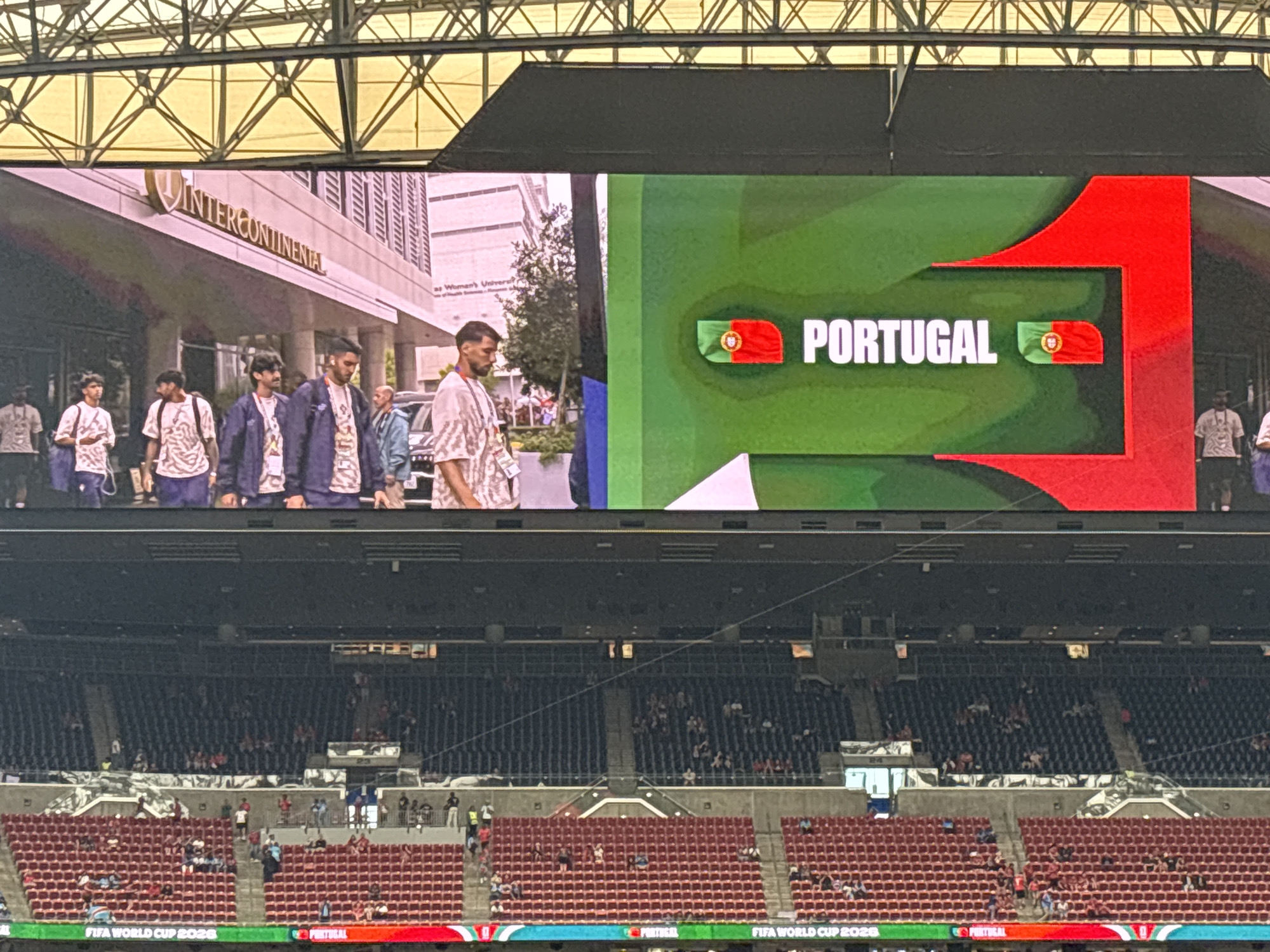 This is a photograph taken at a stadium, likely during a sporting event. The main focus of the image is a large LED screen displaying information related to the FIFA World Cup 2026. The screen shows the word "PORTUGAL" in large white lettering, accompanied by the Portuguese flag. A green and red graphic design frames the text and flag.
Below the screen, rows of empty stadium seating are visible, suggesting that the event has either not yet started or is in its early stages, or that the crowd is sparse. In the background, beyond the screen, a building with the "INTERCONTINENTAL" logo is visible, indicating an urban setting, possibly Houston, Texas, given the context provided.
In the foreground, partially obscured by the stadium structure, are several people. They appear to be walking or standing, and their attire is casual, suggesting they are spectators or staff. The lighting suggests it is daytime. The overall impression is one of anticipation for a major international sporting event. The text "FIFA WORLD CUP 2026" is also visible at the bottom of the screen.