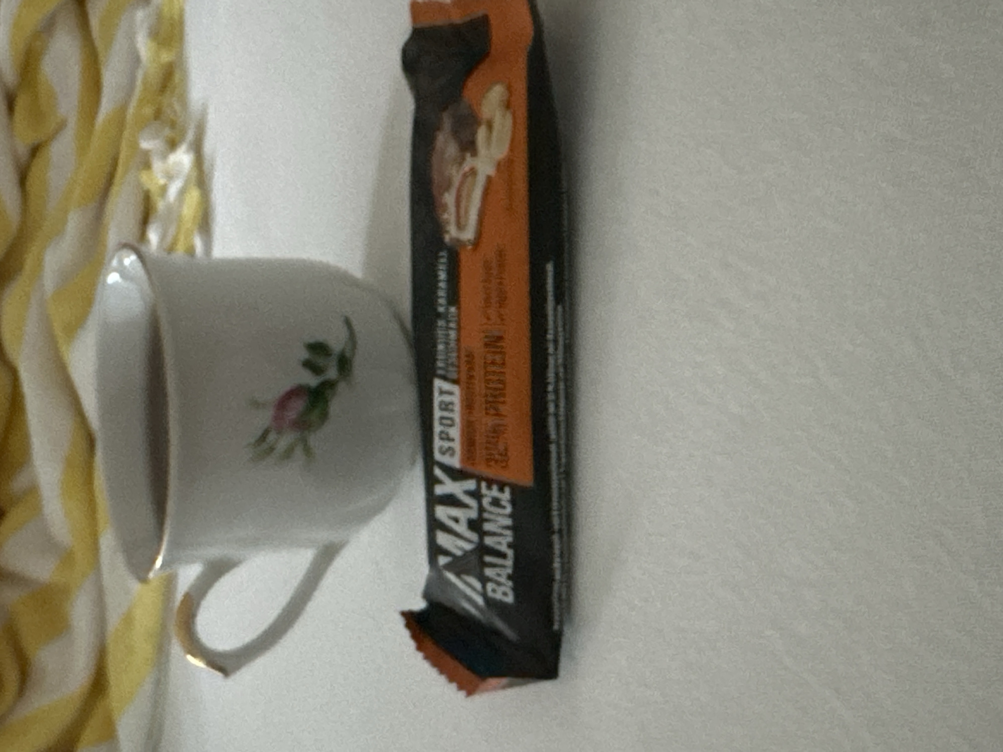 The image shows a white teacup with a gold rim and a pink floral design, filled with a dark liquid. Beside the teacup is a chocolate and caramel protein bar with the branding "MAX BALANCE SPORT". The objects are placed on a white surface, possibly a bedspread. In the background, a yellow and white striped fabric is visible. The scene appears to be indoors, likely in a domestic setting. There is no indication of the location being in Krefeld, Germany.