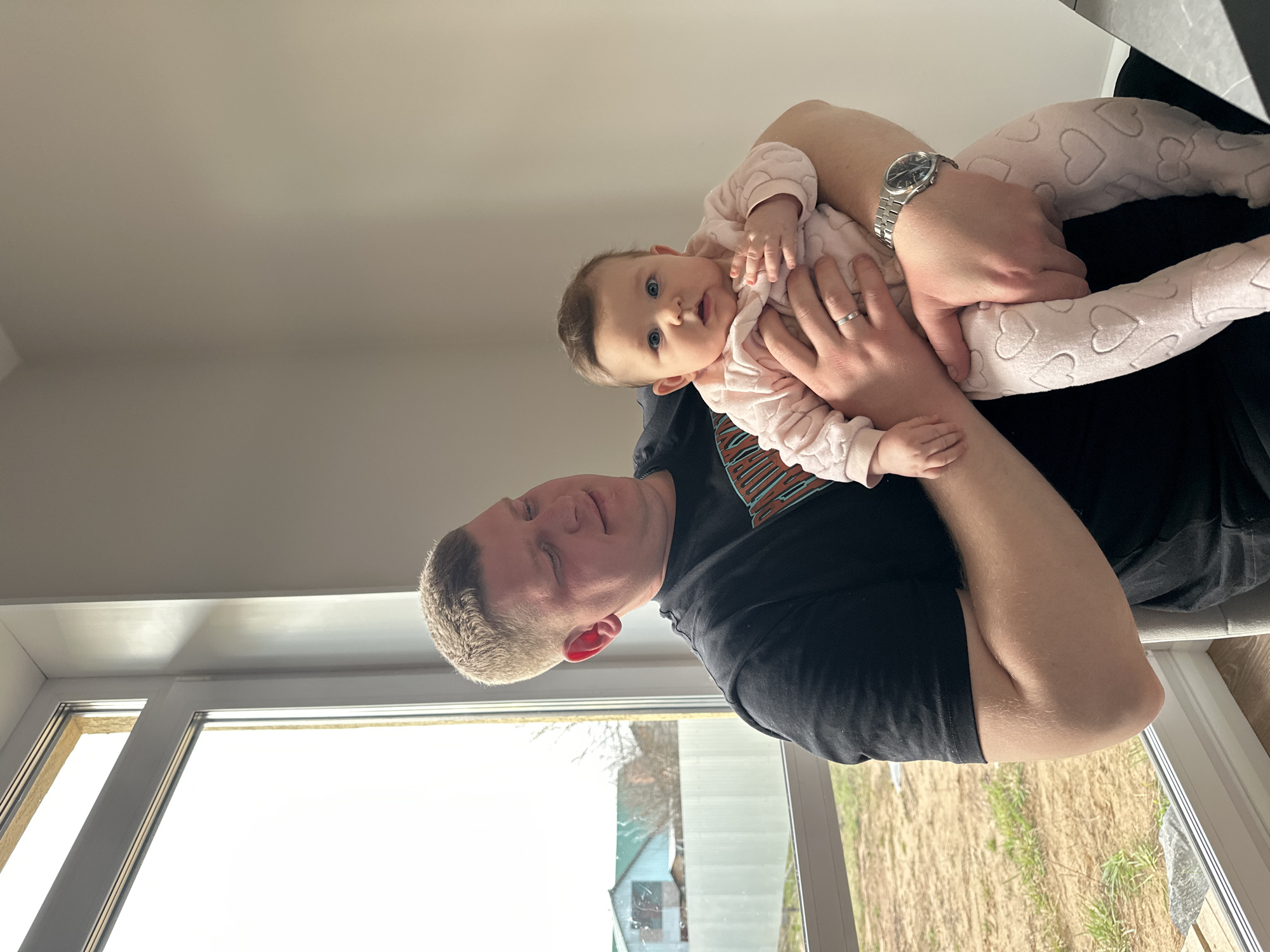 The image shows a man holding a baby indoors, bathed in bright natural light. The man, with short, light-colored hair, wears a black t-shirt with an abstract orange and brown graphic partially visible on his right side. He has a silver-toned watch on his left wrist and a silver ring on his left ring finger. He holds a baby, dressed in a pink, textured outfit adorned with a pattern of hearts, who looks directly towards the viewer with wide, curious blue eyes and slightly parted lips. The baby's small hands are resting on the man's arm.
The setting appears to be a well-lit room, possibly in Chaikovsky, Russia, with a large window on the left side of the frame. Through this window, an outdoor view reveals light-colored ground, sparse green grass, and in the distance, a building with a dark green roof and some bare trees, suggesting a rural or residential environment. The indoor walls and ceiling are a uniform light color. The photograph captures a tender, intimate moment between the man and the baby during daytime hours, highlighting their connection and the baby's curious gaze.
