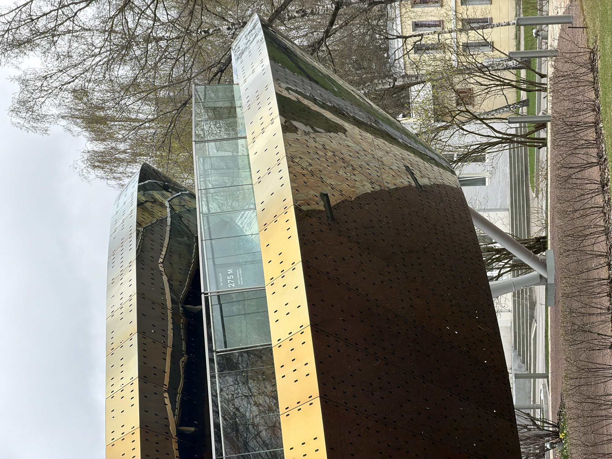 The image depicts a modern architectural structure, likely a building or art installation, against a backdrop of bare trees and a slightly overcast sky. The structure features a reflective, possibly metallic, exterior with a pattern of small rectangular openings or indentations. Adjacent to this section is a glass-encased portion with a sign visible reading "вы прошли 275 м" which translates to "you passed 275 m." This, along with the text mentioning "Мосмос" indicates that this location is possibly at or near the VDNH, an exhibition center in Moscow, Russia. The structure appears to be outdoors, with landscaping and pathways visible in the background. The angle of the photo gives it a disorienting feeling because it is on its side. The image captures the contemporary design of the structure.