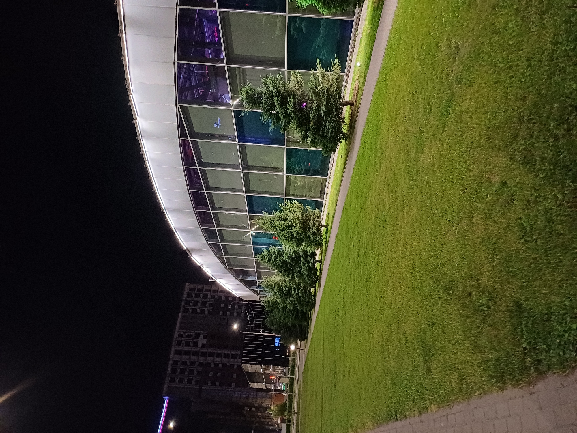 A nighttime, low-angle, exterior, full shot captures a row of fir trees bordering a brightly lit, modern, curved building with large, dark-tinted windows. The building is illuminated from above by a strip of bright white lights along its curved roofline. In the foreground, a neatly manicured green lawn is visible, separated from a paved walkway by a low border. A brick pathway is situated to the left of the lawn. In the background, a dark, unfinished, multi-story building with visible structural elements stands against the night sky. The scene is set in an urban environment, as indicated by the buildings and streetlights, but the specific city and location are unknown. No people or animals are visible in the frame. The lighting suggests it is either dusk or night.