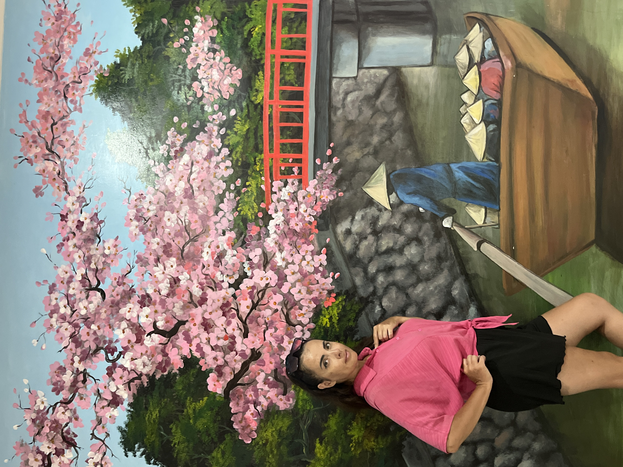 A woman is lying on the ground in front of a mural. The mural depicts a scene with cherry blossoms, trees, a red bridge, and a boat filled with items. The woman is wearing a pink shirt and black shorts. She has dark hair and is wearing sunglasses on her head. The mural appears to be in a garden setting. The time of day is not discernible, nor is the weather. The location is Kuta Selatan, Indonesia.