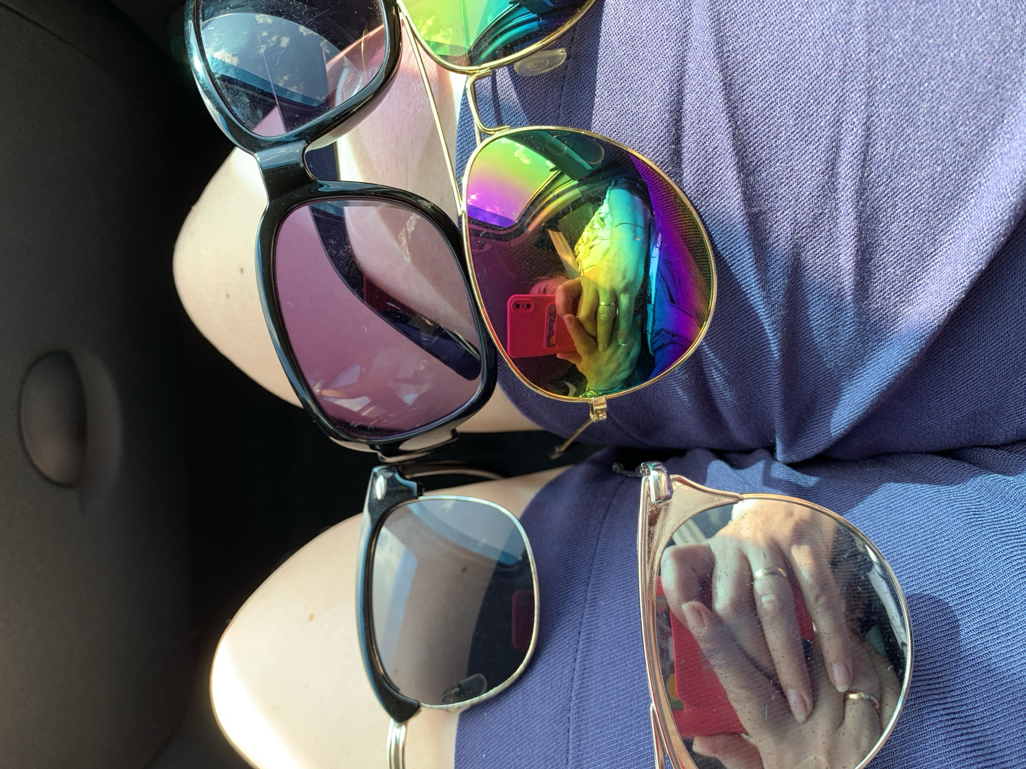 The image depicts a close-up view of a person's lap, covered by a textured blue fabric, likely clothing. Several pairs of sunglasses are arranged on the fabric. In the foreground, a pair of gold-rimmed sunglasses with mirrored, rainbow-colored lenses dominates the view. The reflection in these lenses shows a person holding a red smartphone and wearing rings on their fingers. Behind these, to the left, is a pair of black-rimmed sunglasses with dark lenses. To the right, another pair of black-rimmed sunglasses with purple-tinted lenses is visible. The background is dark and out of focus, suggesting an indoor or shadowed environment, possibly the interior of a vehicle. The image does not provide information about the specific location within Азерскі сельскі Савет, Belarus.