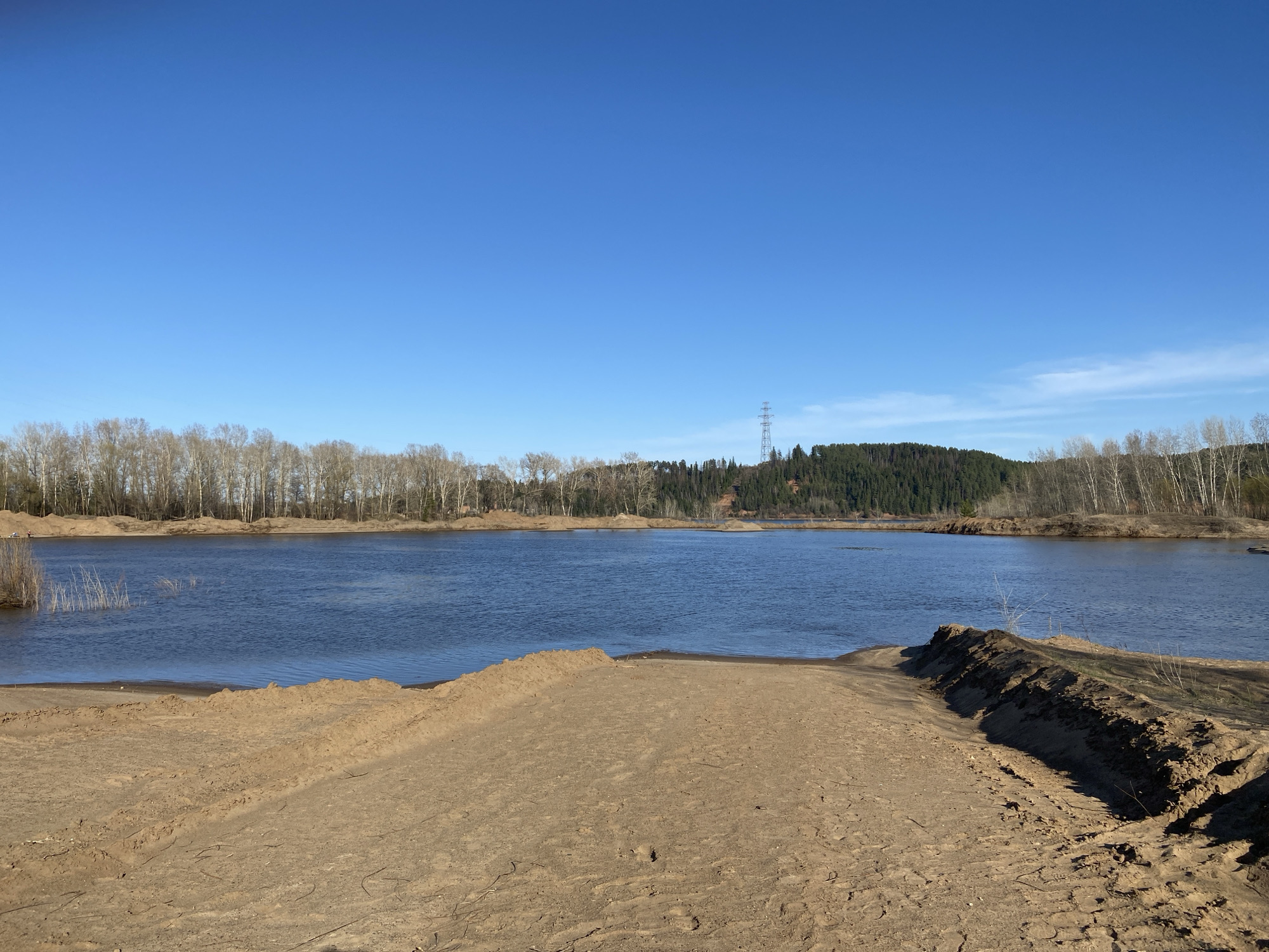 The image shows a sandy area leading to a body of water under a clear blue sky. Across the water, a treeline is visible, with a forest patch on the right side of the scene. An electrical transmission tower is visible in the distance. The location is Noviy, Russia. There are no people visible in the frame. The environment is characterized by sand, water, and trees. The season appears to be either late fall or early spring, due to the bare trees.