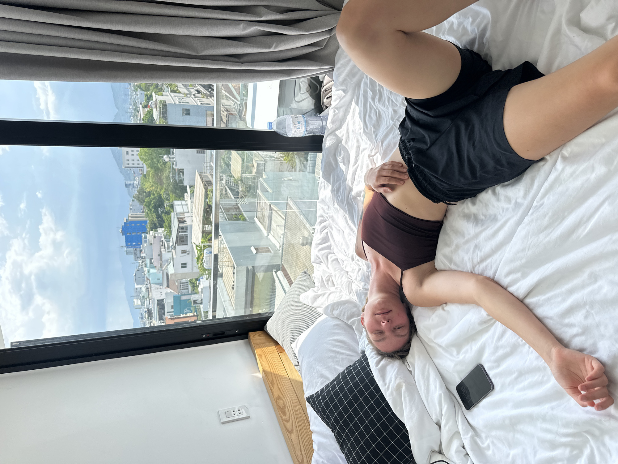 A young Caucasian woman is lying on a bed covered with white sheets, her body angled slightly to the right. She is wearing a dark red tank top and black shorts. Her eyes are closed, and her head is resting on a pillow. A smartphone is placed on the bed to her left. Behind her, a large window overlooks a city scene in Đà Nẵng, Vietnam. Buildings of various colors and sizes are visible, along with trees and mountains in the distance under a cloudy sky. A gray curtain hangs on the right side of the window. A partially filled bottle of water is also on the bed near the window. The scene suggests a moment of rest or relaxation.