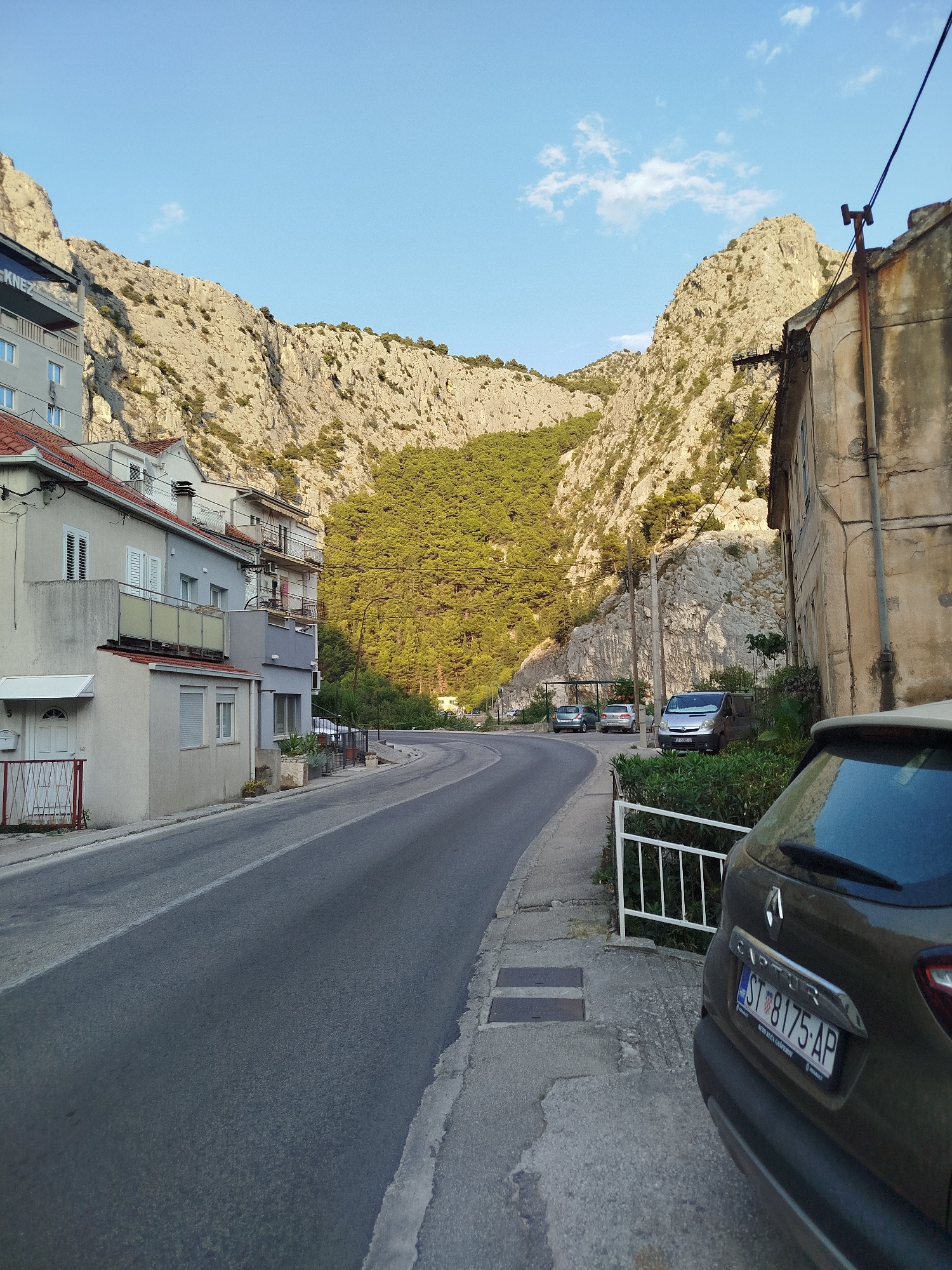 The image shows a street in Omiš, Croatia, with steep, rocky mountains and dense green trees in the background. The street curves to the left, lined with multi-story buildings on the left and a weathered, concrete building on the right. Several cars are parked along the street. A dark-colored Renault Captur is parked in the foreground on the right, its rear facing the camera, and its license plate is visible. The sky is clear blue with a few scattered clouds. The lighting suggests it is late afternoon or early evening, with shadows cast by the buildings and mountains. The overall scene depicts a somewhat urban environment nestled within a dramatic natural landscape.