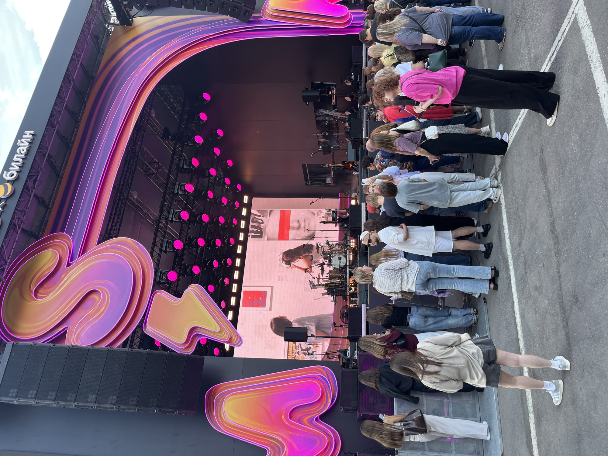 The image depicts an outdoor stage setup at a public event, likely a concert or festival, at the Khimki Arena in Khimki, Russia. The stage features a large, illuminated screen displaying a performance, with prominent, stylized letters forming the word "MTS" in bright pink and orange neon-like lights on either side of the screen. Above the screen, the logo of "Beeline" is visible, suggesting sponsorship.
A crowd of spectators, diverse in age and gender, stands in front of the stage, facing the screen. They are dressed in casual attire, typical for an outdoor event. Some are standing, while others appear to be interacting with each other or looking at their phones. The ground is paved, likely asphalt, with white lines marking areas. The overall atmosphere suggests a lively and energetic event.