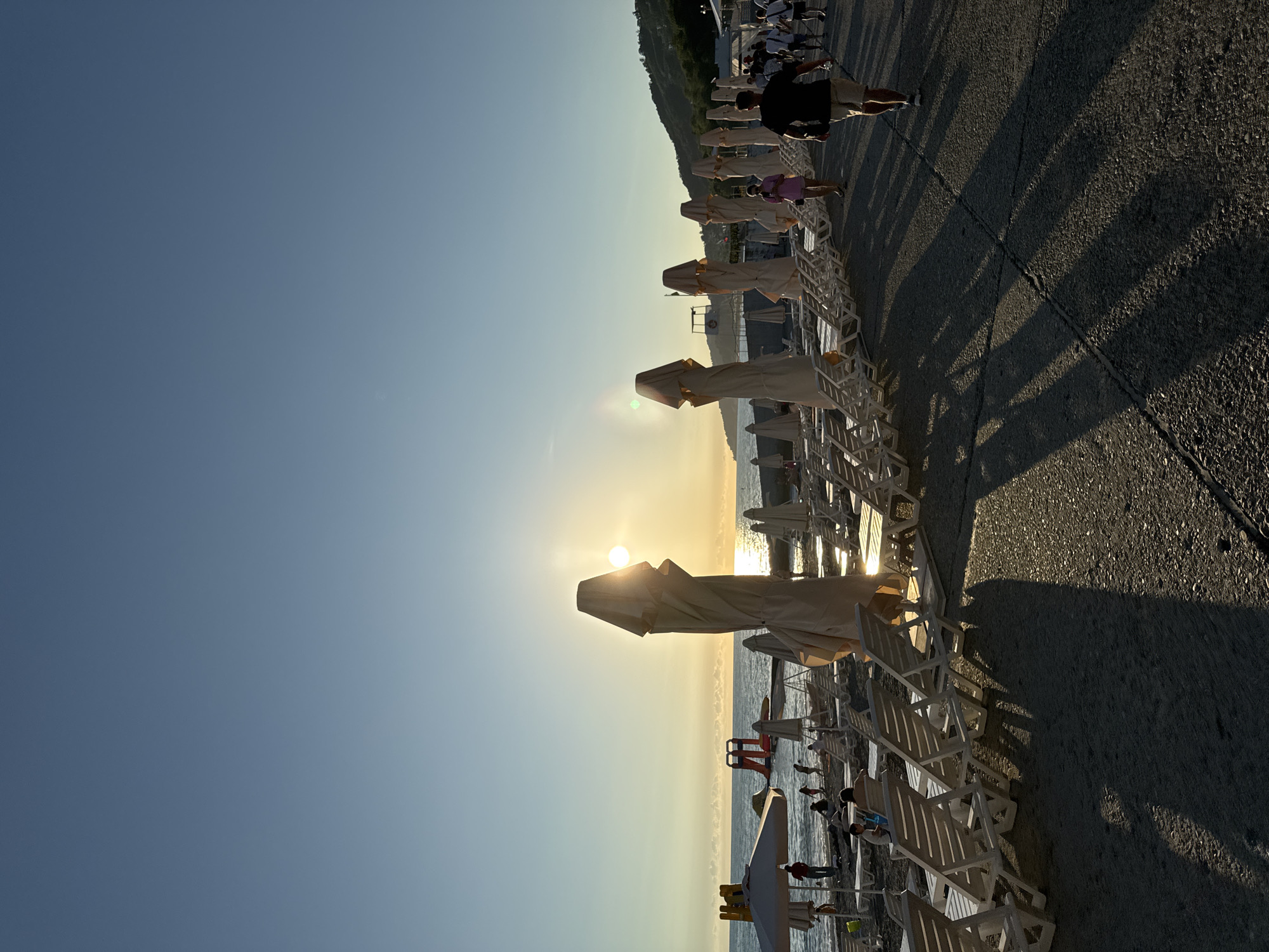 The image depicts a beach scene in Sochi, Russia, during sunset. The sun is low on the horizon, casting a warm golden light across the water and the beach. In the foreground, rows of empty lounge chairs and closed umbrellas are arranged on a paved walkway. Several people are walking along the walkway, casting long shadows on the ground. In the background, the sea is visible, with a few people on the beach and a water slide. The sky is clear and blue, with a gradient towards a lighter hue near the horizon. The overall atmosphere is peaceful and serene, capturing the end of a day at the beach.