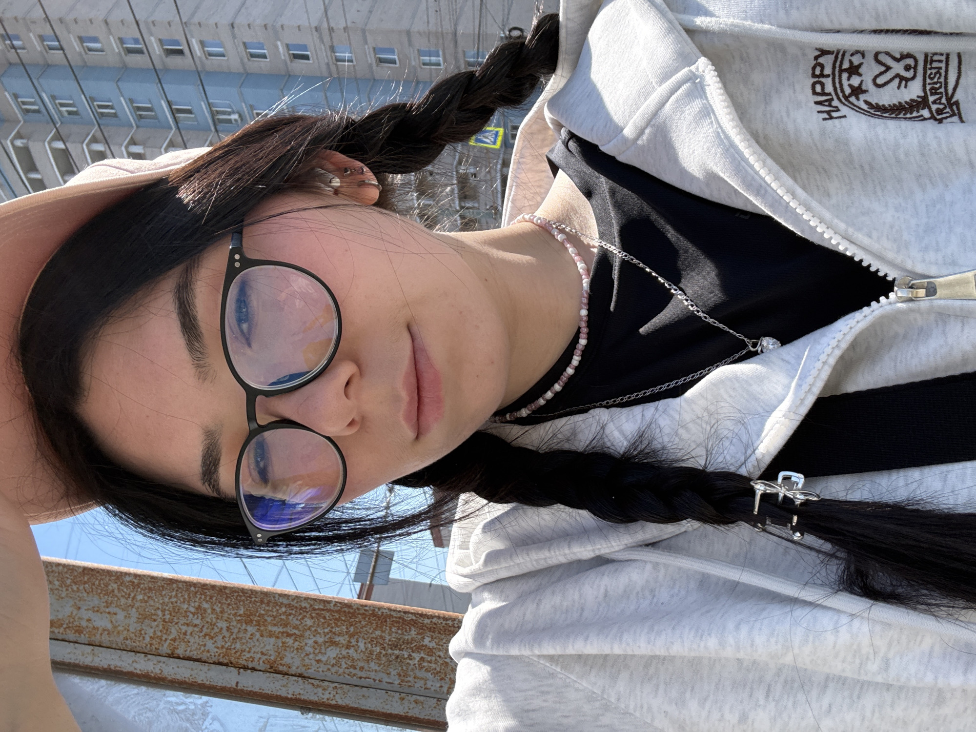 A woman with long black hair braided into pigtails and glasses is looking at the camera from an elevated point in Izhevsk, Russia. She is wearing a black t-shirt and a grey zip-up hoodie with a logo on the chest. She also wears a pink and white beaded necklace and a silver chain. A metal railing is visible in the foreground. In the background is a large, multi-story residential building with numerous windows. The sky is light blue.
