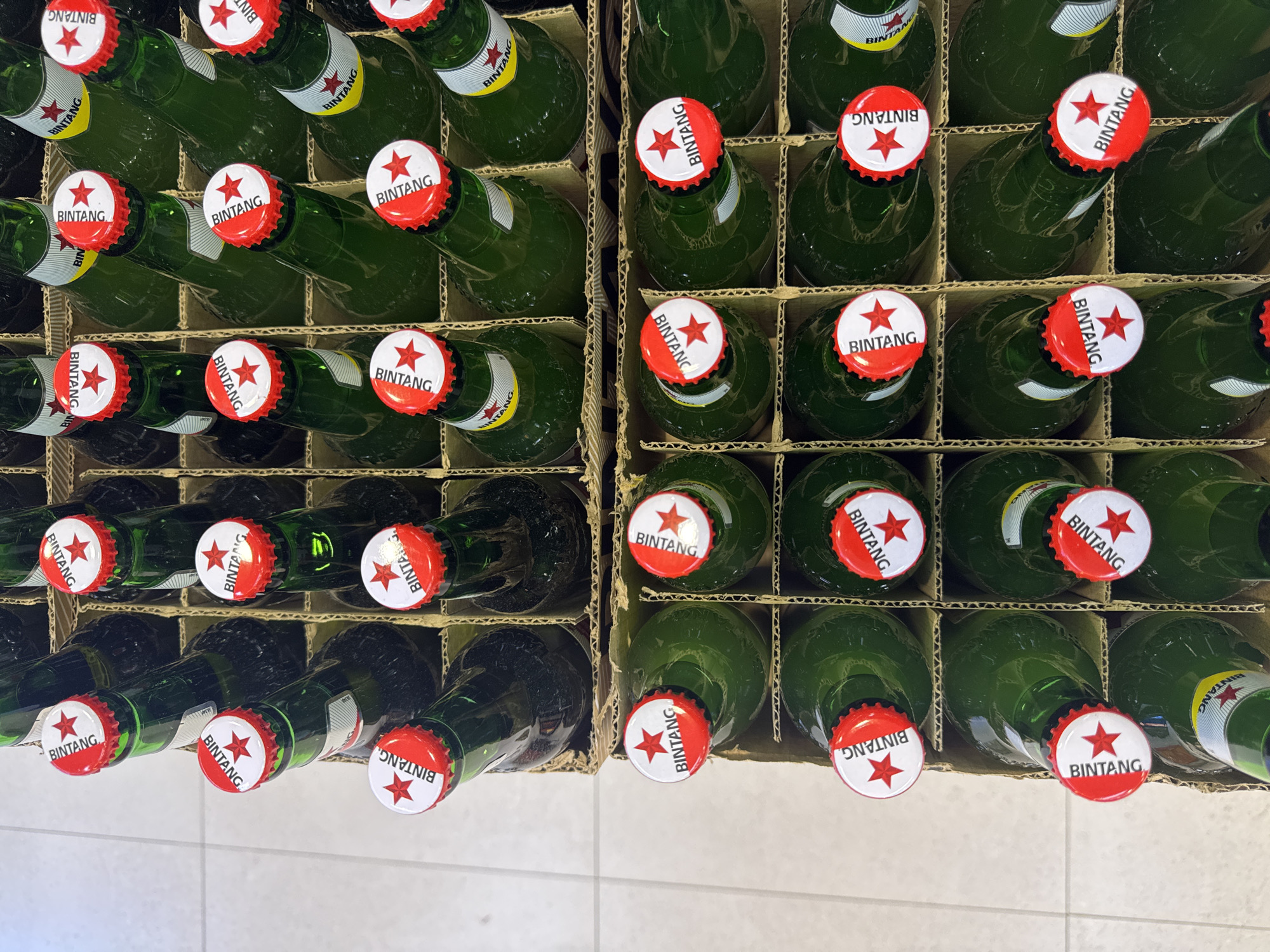 An overhead view captures numerous green glass bottles, likely containing beer, neatly packed in brown cardboard crates. The bottles are arranged in a grid-like pattern, with some standing upright and others slightly angled. Each bottle is sealed with a distinctive red and white crown cap that features a red star and the word "BINTANG" printed in white. On several caps, the "BINTANG" text is visible, sometimes appearing inverted depending on the bottle's orientation. The bottles also have labels, some of which clearly display "BINTANG." The cardboard dividers segment the crates, suggesting a storage or retail display arrangement. A light-colored tiled floor is visible at the bottom, indicating an indoor setting such as a store or warehouse in Kuta Selatan, Indonesia.