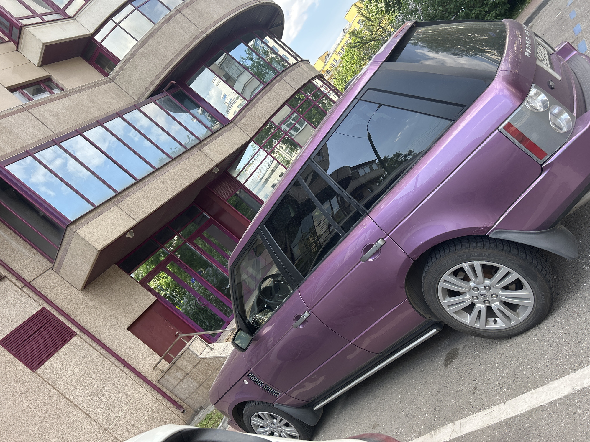 The image shows a purple Range Rover parked at an angle in front of a modern building. The building has a facade of beige and purple, with large glass windows reflecting the sky. The car is parked on a paved surface, with a white parking line visible. The tires of the car are silver and black. The location is in the city of Unknown, Unknown.