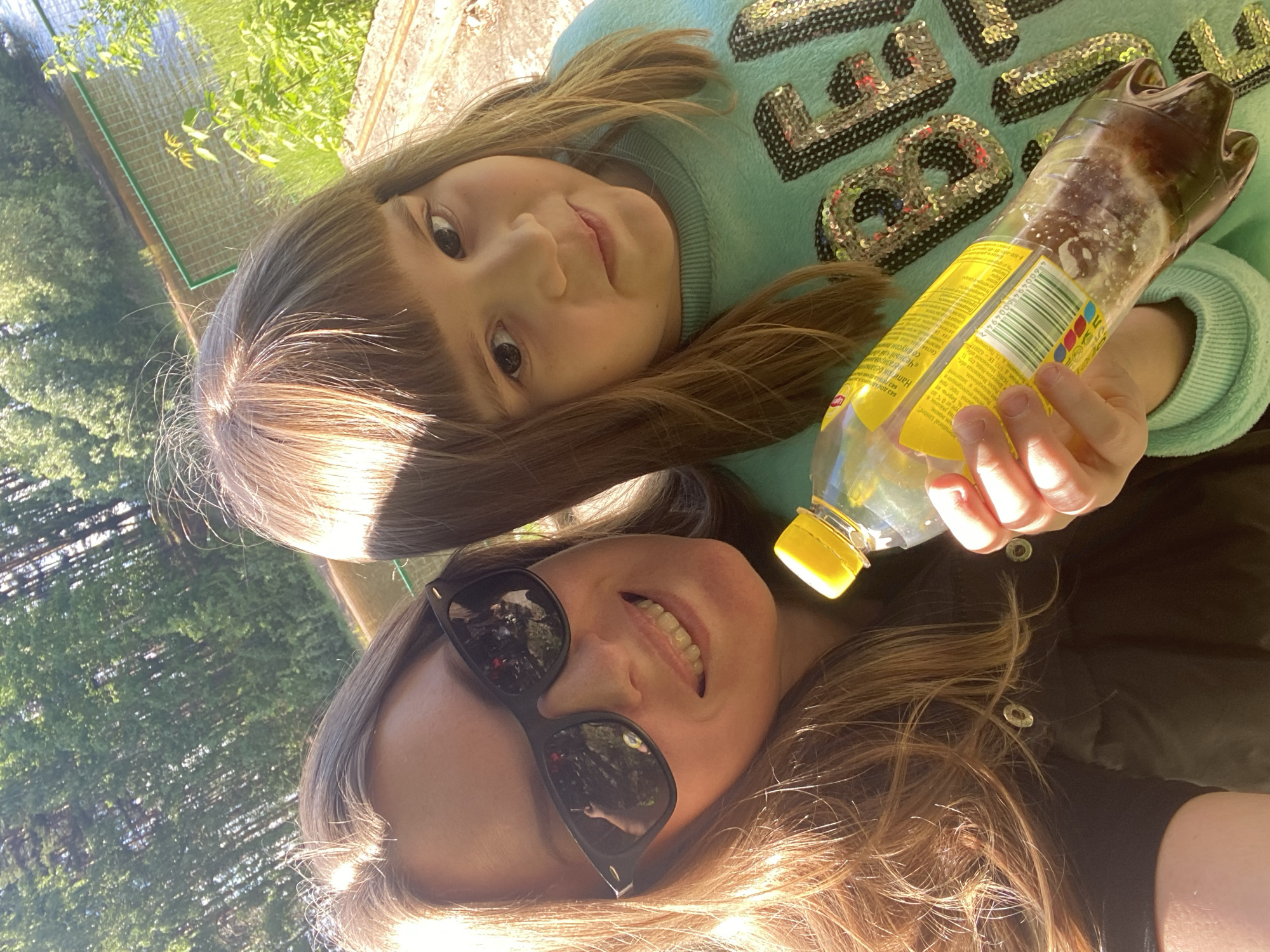 A selfie-style shot captures two individuals against an outdoor backdrop, likely near a body of water. The foreground features an adult wearing sunglasses and a dark-colored garment, while a child in a light blue sweatshirt with sequined letters appears above and slightly behind. The child has light brown hair with bangs and is looking at the camera. The adult also gazes at the camera, smiling. A plastic bottle with a yellow label is held in the hand of the child. The bottle's label appears to have Russian text on it. The background includes greenery and what appears to be water and a fence. Based on the OCR content on the bottle, the product is labeled and packed in Russia, possibly in the Moscow region. The product is labeled to be a non-carbonated black tea with an unspecified taste. It is unclear what the specific city is, but one EK-620017 address is associated with Chaikovsky, Russia.
