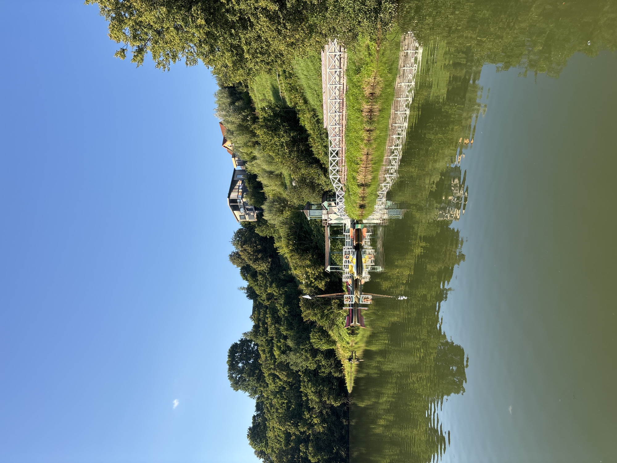 The image depicts a serene lakefront scene in Romashkovo, Russia, under a clear blue sky. On the left side, the tranquil green water of the lake reflects the lush, tree-covered hillside that slopes upwards. Several small boats, including what appears to be a red rowboat and yellow paddle boats, are docked near a wooden pier with a white railing. A small pavilion structure sits on the pier, providing shelter. The hillside is densely wooded with various trees, and in the distance, two modern houses are visible nestled on the higher ground. The water surface is calm, with gentle ripples and clear reflections of the surrounding greenery and structures. The overall impression is one of a peaceful natural setting.
