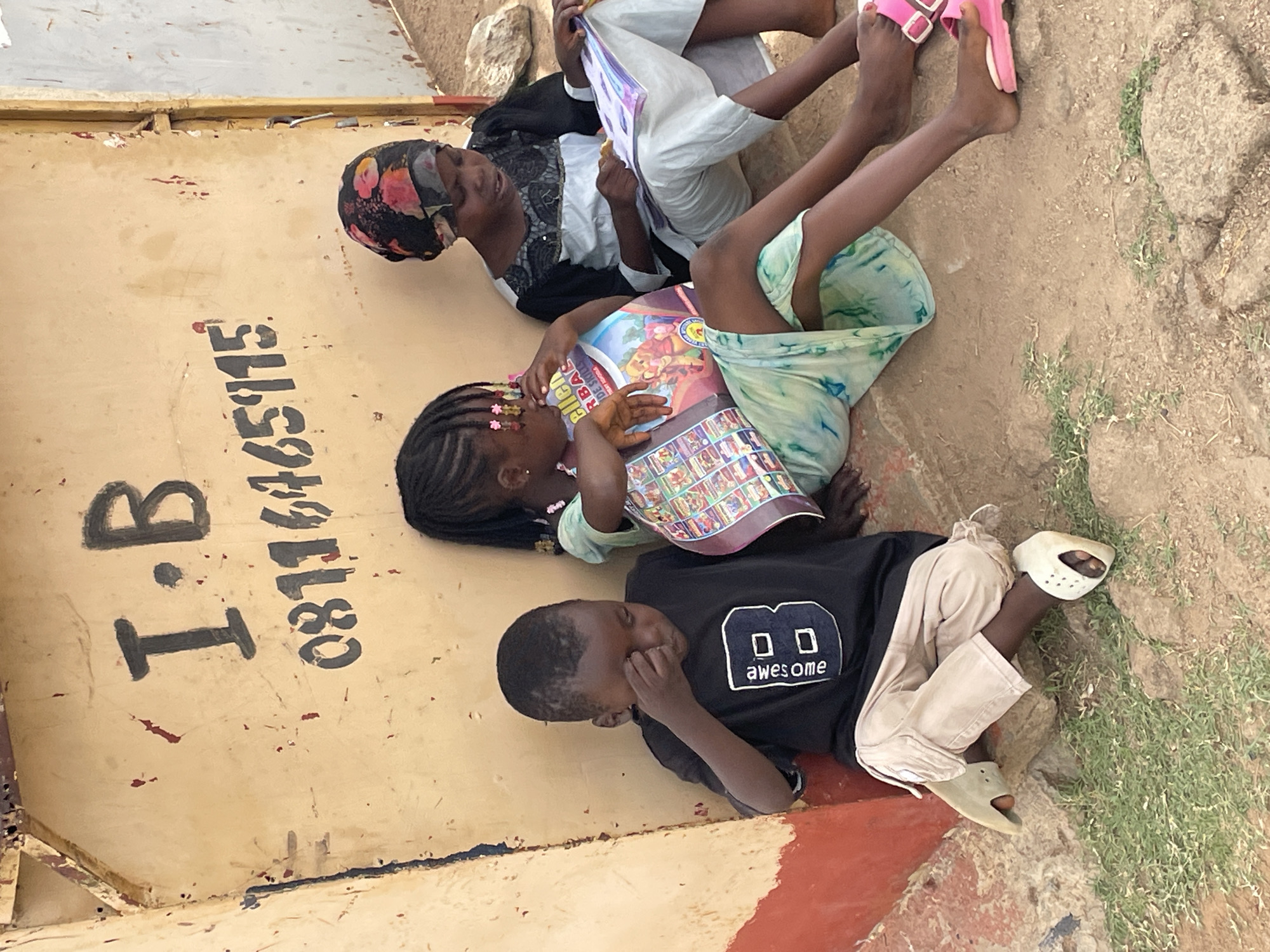Three children are lying on the ground next to a yellow wall with black lettering that reads "I.B. 08116465115". The children are oriented at various angles, their bodies partly overlapping. The child closest to the wall is wearing a black t-shirt with a white design reading "awesome" and tan-colored pants. This child has their right hand covering the lower part of their face. The middle child is wearing a light green patterned dress and is holding a colorful book or magazine. The furthest child is wearing a black and white patterned head covering and dress, and white pants. This child is also holding a book. The ground around the children is dirt and sparse grass. The location is Jos, Nigeria.