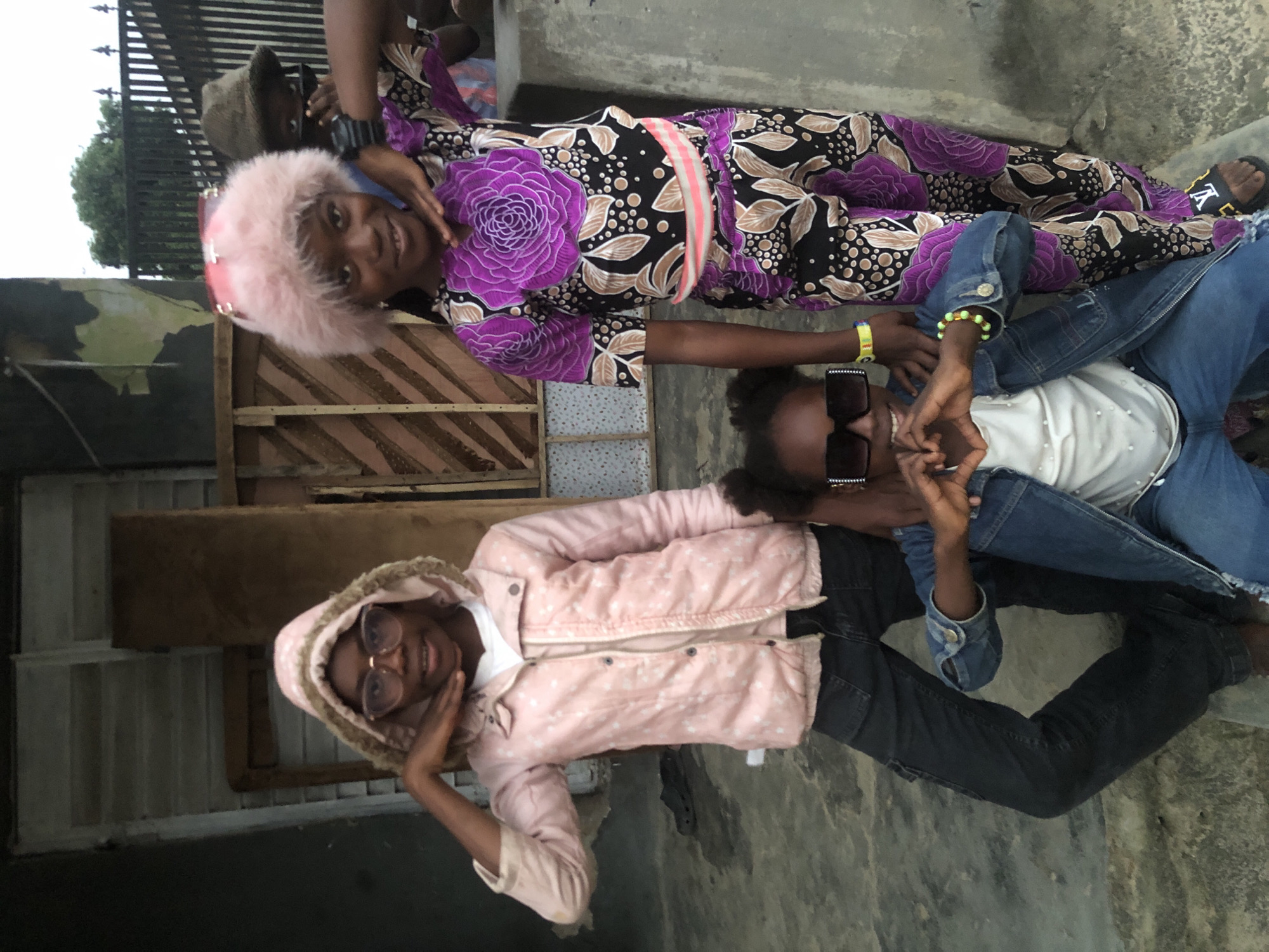 Three young girls are posing for a photograph outdoors in a residential area, likely Ado, Nigeria. The girl on the left wears a pink hooded jacket with white polka dots, a white t-shirt, and dark jeans. She has glasses and is touching her chin. The middle girl is seated and wears a denim jacket over a white t-shirt and jeans. She has large square sunglasses and is forming a heart shape with her hands. The girl on the right is standing and wears a purple and black patterned outfit with a large floral design on the chest and a pink sash. She has a fluffy pink hat on her head and is touching her face. In the background, there is a wooden structure with vertical planks and a metal fence. The ground is concrete.