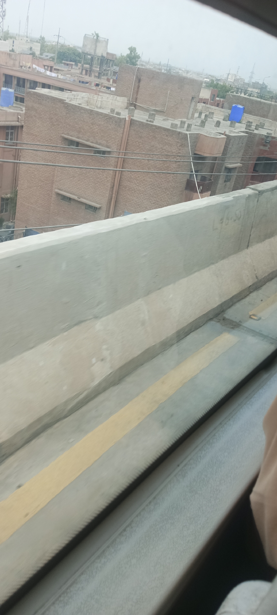 The image shows an elevated view of a city, likely Alam Khan Kalay, Pakistan, from the perspective of a moving vehicle. In the foreground, a concrete barrier with a yellow stripe runs diagonally across the frame. The background features several multi-story brick buildings with visible windows and some with blue water tanks on their roofs. The sky appears overcast. There are some visible electrical wires and a hand is partially visible on the right side of the frame, suggesting the photo was taken from inside a vehicle. The overall scene depicts an urban environment with a slightly hazy atmosphere. A handwritten inscription, "L 46-50", is visible on the concrete barrier.