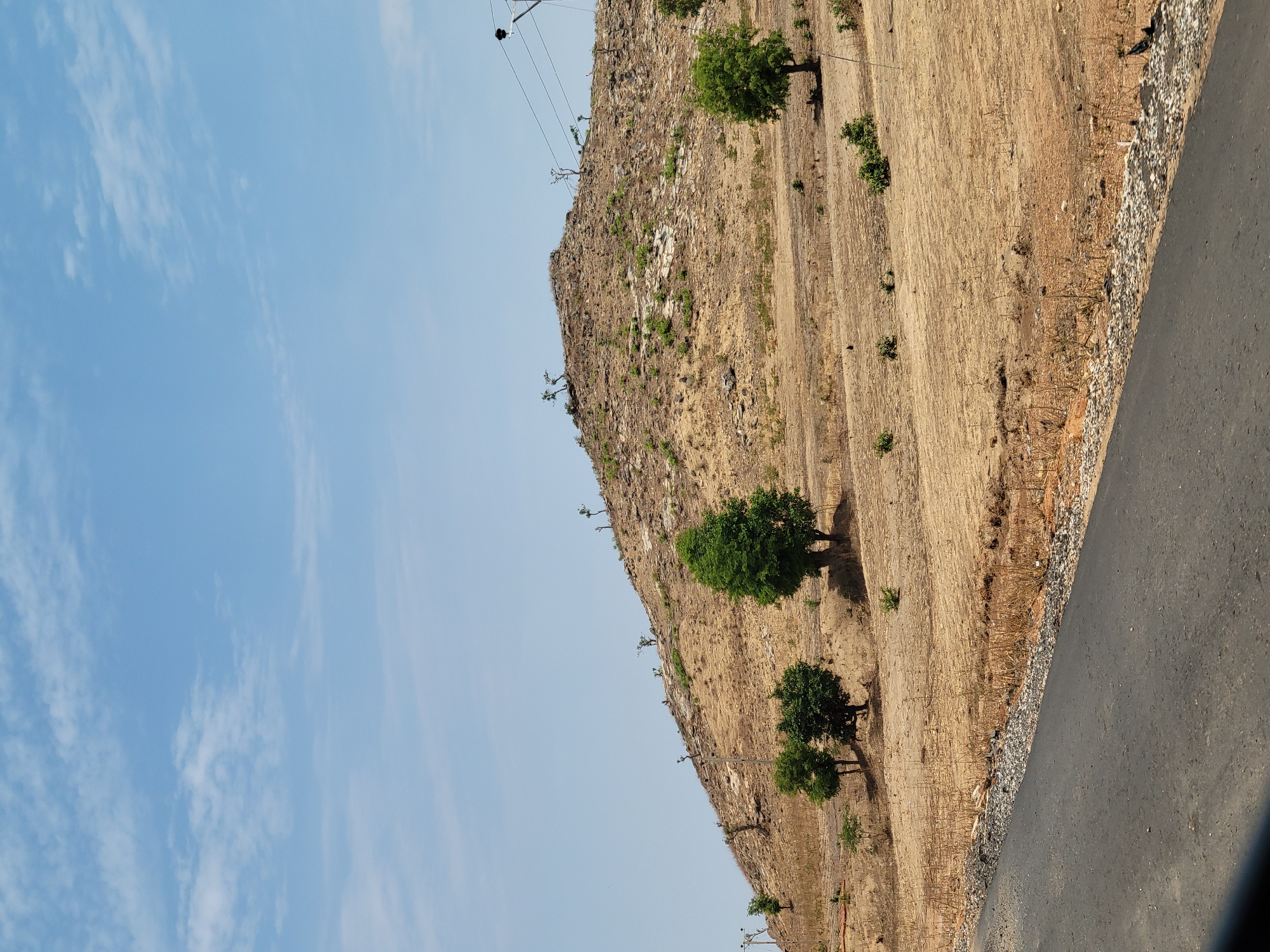 The image shows a roadside view of a dry, hilly landscape in Jama'a, Nigeria. The hillside is sparsely vegetated with brown grass and scattered trees. A paved road runs along the base of the hill. Power lines are visible along the top of the hill. The sky is light blue with some clouds.