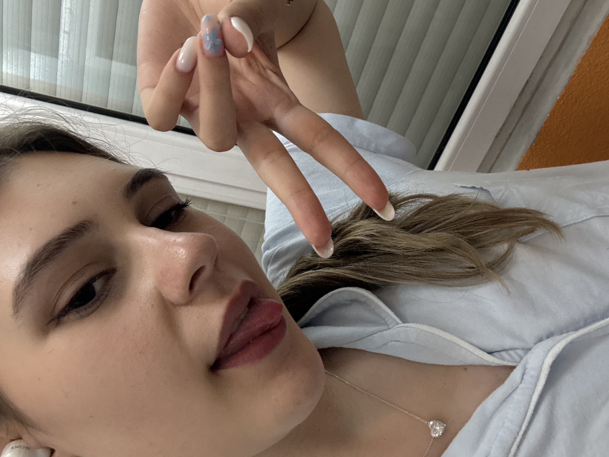 A close-up, angled shot depicts a woman lying down, her face tilted towards the camera. She has light skin, dark eyebrows, and dark eyes. Her lips are slightly parted, revealing a hint of her tongue. She is wearing lipstick. Her hair is blonde, with strands visible near her neck and shoulder. A silver necklace with a heart-shaped pendant is visible on her chest. Her left hand is raised, fingers spread in a "peace" or "victory" sign. The fingernails are painted with a white base and a blue flower design on the thumb. The background consists of a window with vertical blinds and a segment of a wall painted in orange. There is no information to confirm the location is in Panagyurishte, Bulgaria.