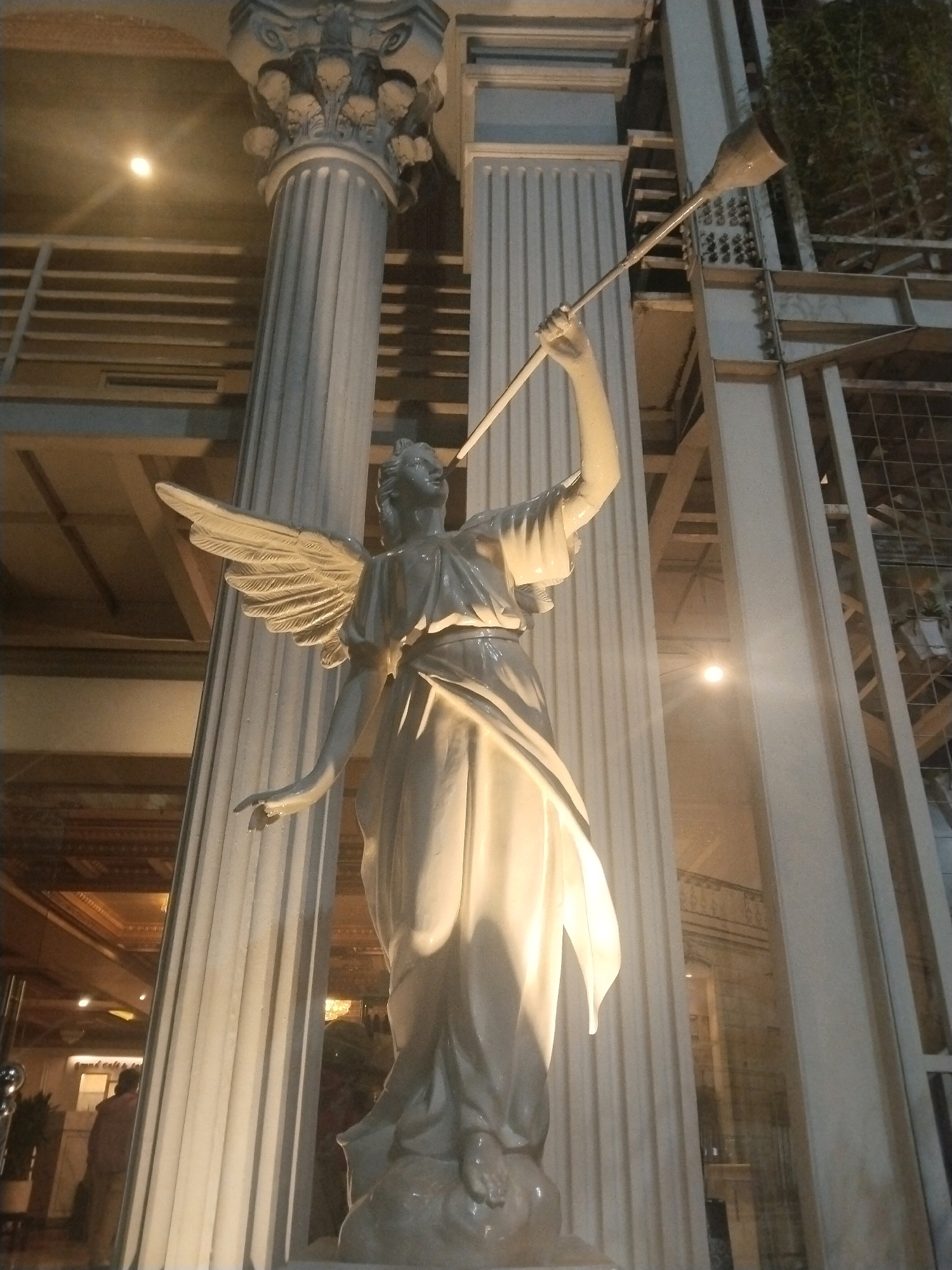 A low-angle, full shot captures a white, angelic statue with wings and a trumpet, positioned between two fluted columns. The statue is illuminated by ambient light, creating strong shadows and highlights. In the background, the interior of a building is visible, with a person standing near a doorway and illuminated by a bright light. The architecture suggests a grand, possibly indoor space with decorative elements and metal structures. The scene appears to be within a commercial or public building in Sinduadi, Indonesia, though the specific location is not explicitly identified.