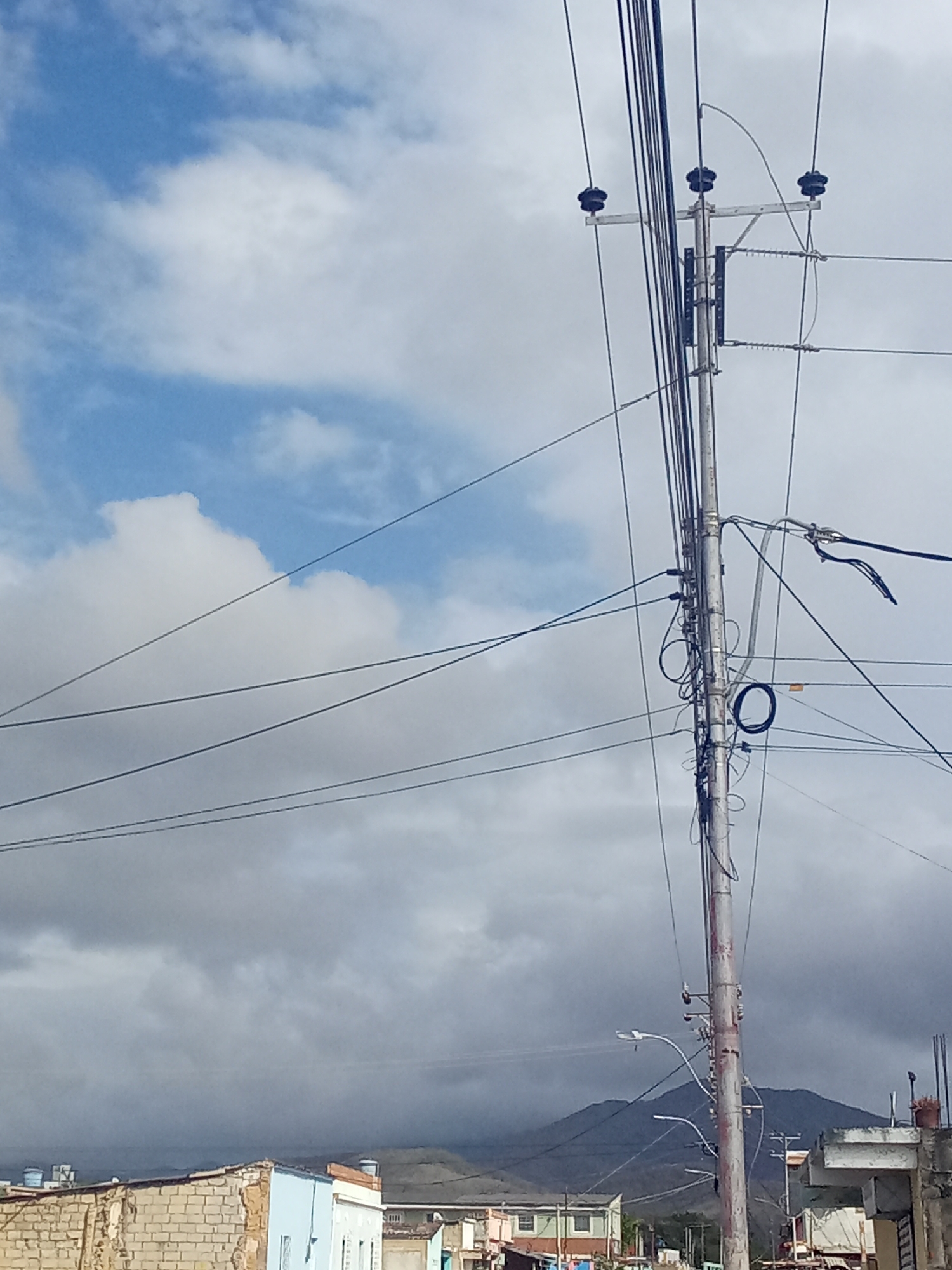 The image depicts a scene in Juan Griego, Venezuela. The primary focus is a utility pole laden with electrical wires and communication cables. The pole is positioned against a backdrop of partly cloudy sky and overcast weather. Below the horizon line are buildings, some under construction, and hints of a hillside or mountain in the distance. There are no people visible in the image. The dominant objects are the utility pole, wires, cables, buildings, and sky. The interactions are implied by the presence of electrical and communication infrastructure, suggesting energy distribution and connectivity within the town. The environment is characterized by a mix of urban development and natural landscape under a cloudy sky.