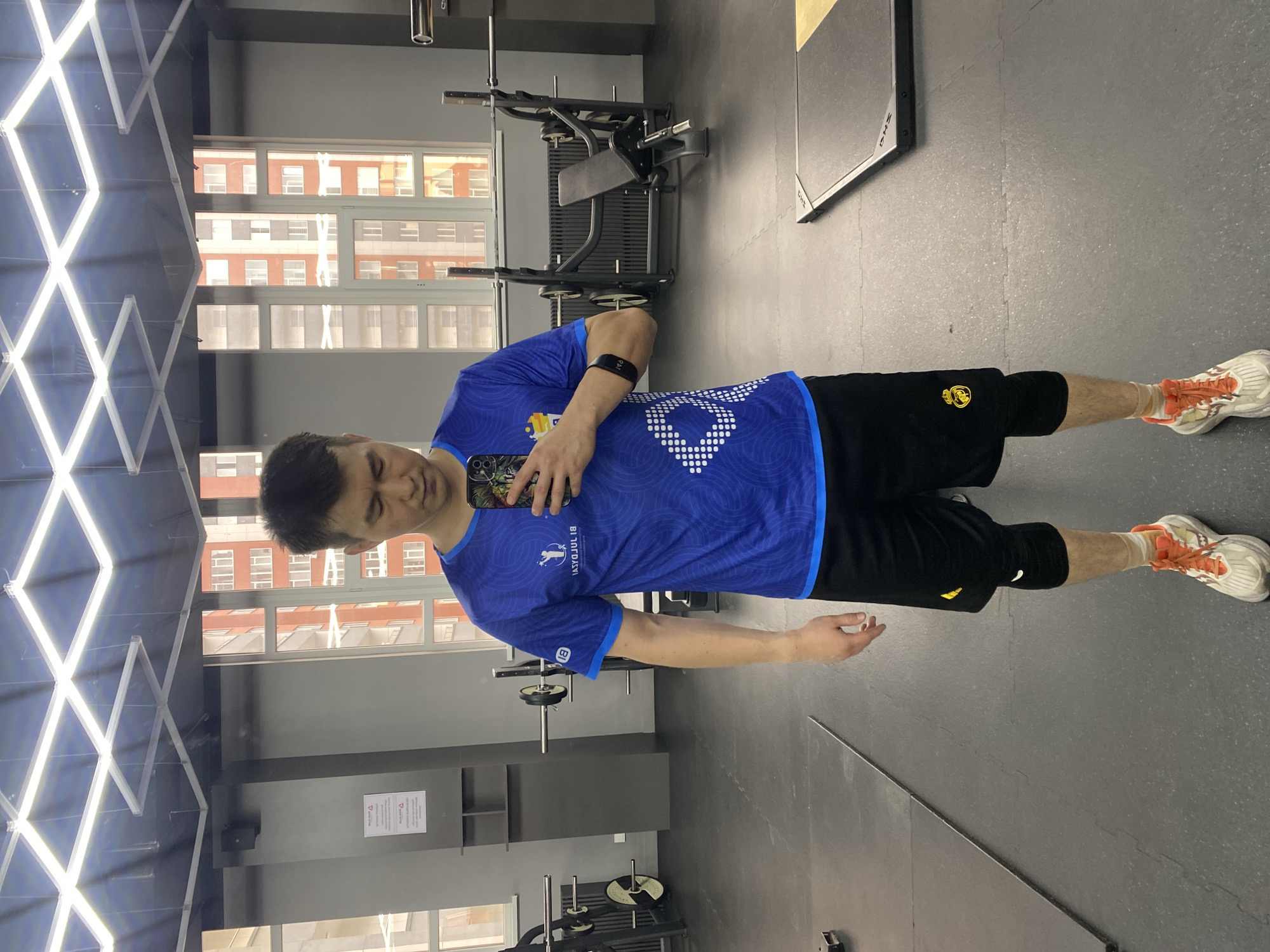 A full-body, front-facing mirror selfie of a man in a gym located in Astana, Kazakhstan. The man is wearing a blue t-shirt with white circular patterns and a logo on the left chest, black shorts with a yellow emblem on the left thigh, knee pads, and white and orange athletic shoes. He is holding a smartphone in his right hand. The gym has a dark gray floor, black walls, and large windows overlooking buildings. There are weightlifting racks, benches, and other gym equipment visible in the background. The lighting appears to be fluorescent overhead lights.