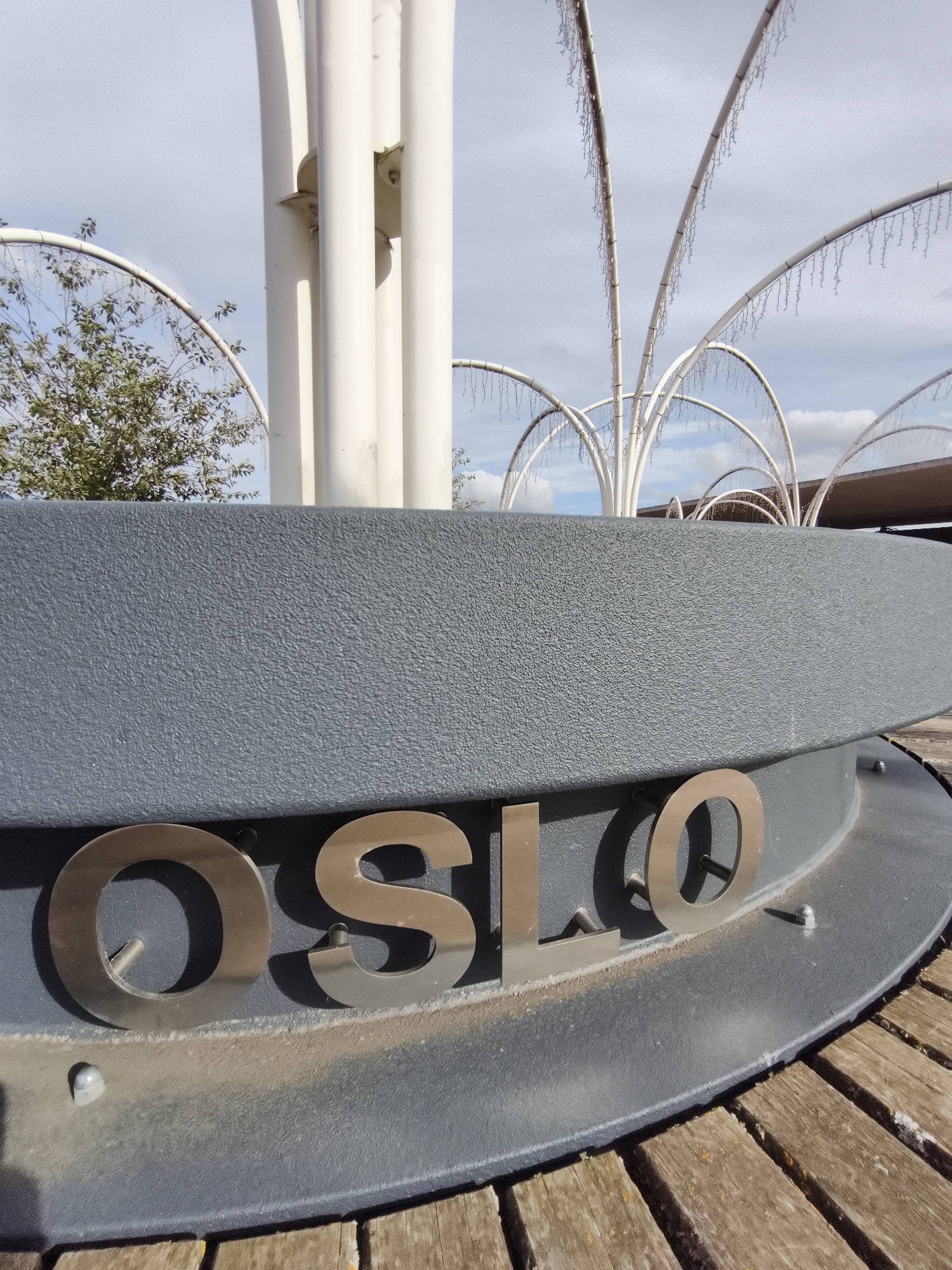 The image shows a close-up view of the word "OSLO" spelled out in large, metallic letters attached to a dark grey, textured curved structure. The letters are silver and appear to be three-dimensional. Behind the curved structure, there are white, arched elements adorned with what seem to be small lights or decorations, creating an elaborate canopy-like effect against a cloudy sky. To the left of the central structure, a portion of a green tree with leaves is visible. The foreground features wooden planks, suggesting an outdoor seating area or pathway. The scene is well-lit, indicating it was taken during daylight hours. No people are present in the image. The location is not specified as Tallinn, Estonia.