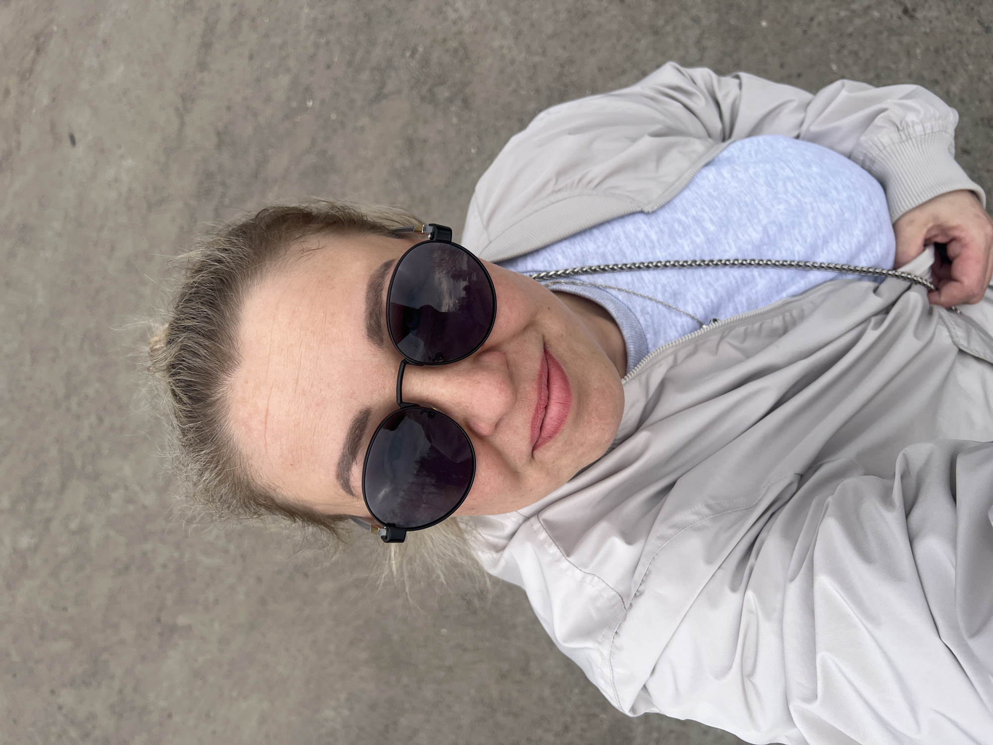 A person wearing round sunglasses and a light gray jacket takes a selfie in Chaikovsky, Russia. The ground is light gray concrete. The person is wearing a light blue shirt under the jacket and has a silver chain visible around their neck. The sunglasses reflect buildings and trees, suggesting an urban environment. The person is smiling slightly. The overall tone of the photo is casual and relaxed.