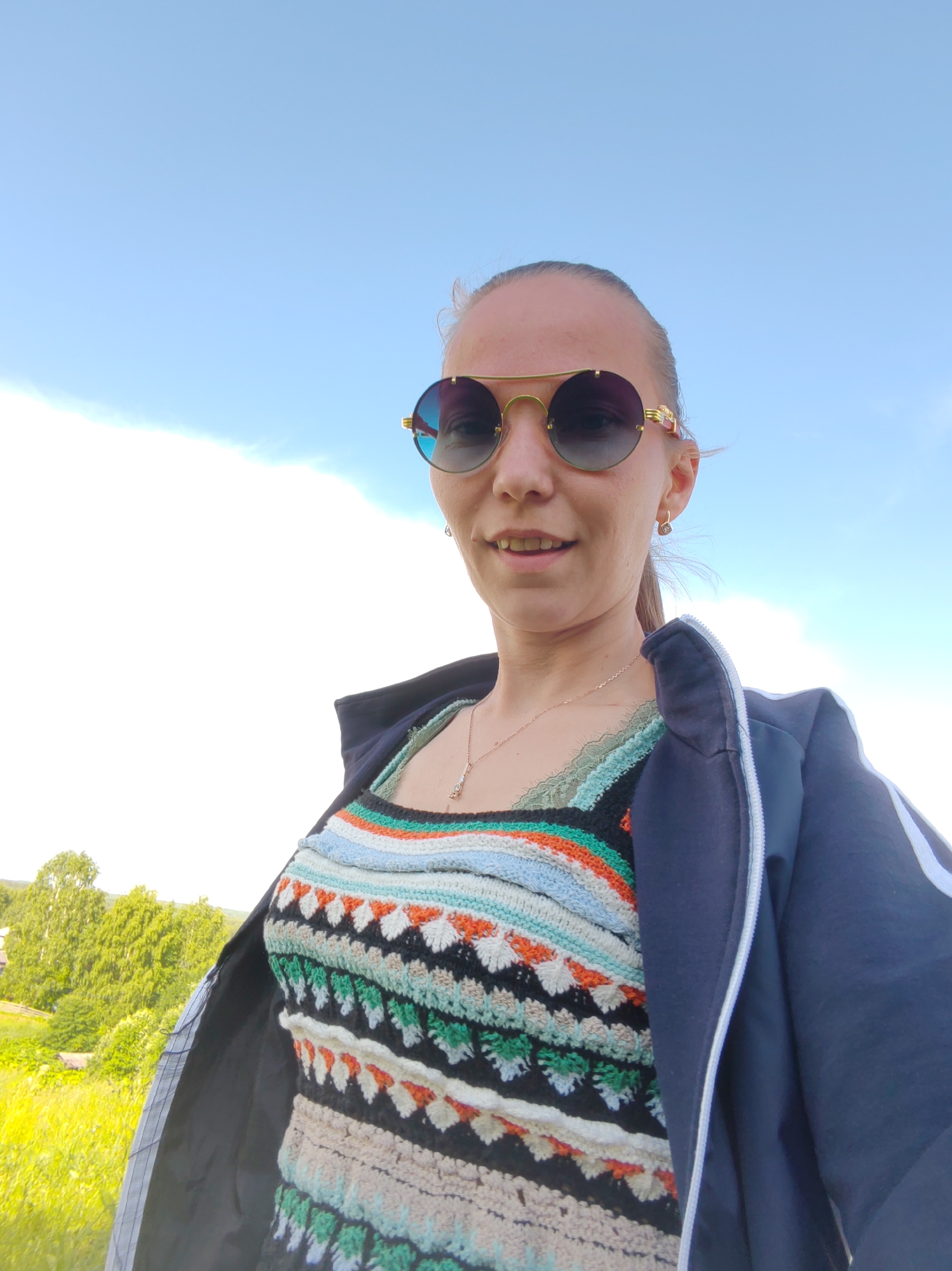 A woman is captured in a selfie taken outdoors under a bright blue sky with scattered clouds. She is wearing round, gold-rimmed sunglasses with gradient lenses, and a dark blue jacket partially unzipped over a colorful, crocheted tank top. The tank top features a geometric pattern with horizontal stripes in shades of blue, white, orange, green, and black. A delicate gold necklace with a small pendant is visible around her neck. Her hair is pulled back, and she has a subtle smile on her face, suggesting a pleasant and relaxed mood.
The background reveals a vibrant green landscape with rolling hills and trees, indicative of a rural or natural setting. The bright sunlight and clear sky suggest it is daytime, likely during the warmer months. The overall impression is that of a casual, outdoor moment, possibly during a trip or leisurely activity.
Given the location context of Чухлэм, Russia, the scene likely depicts a summer day in the Russian countryside. There is no visible text in the image.