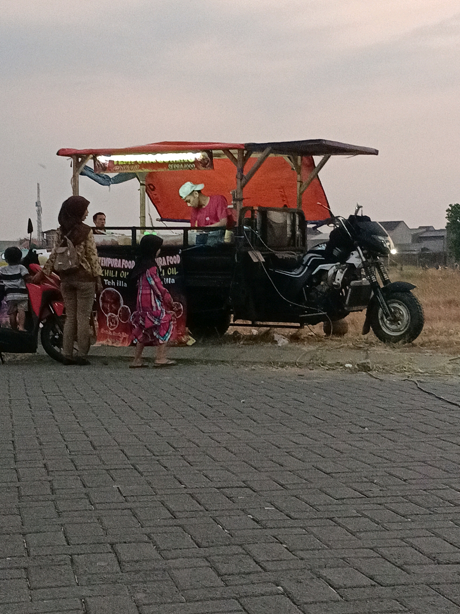 A three-wheeled motorcycle, modified with a cart and an overhead awning, is parked on a paved surface. The cart appears to be a food stall, with an orange tarp covering the top and a white sign with text. A person wearing a pink shirt and a white head covering is visible behind the counter, seemingly serving customers. In front of the stall, two women and a young child are interacting. One woman, wearing a headscarf and a patterned backpack, stands beside a red scooter. Another young girl, dressed in a colorful pink and blue dress, walks away from the stall. A second man is partially visible inside the cart. The background shows a grassy field with some structures and trees, suggesting a suburban or rural setting. The sky is overcast. The overall scene depicts a street food vendor operating in Kletek, Indonesia.