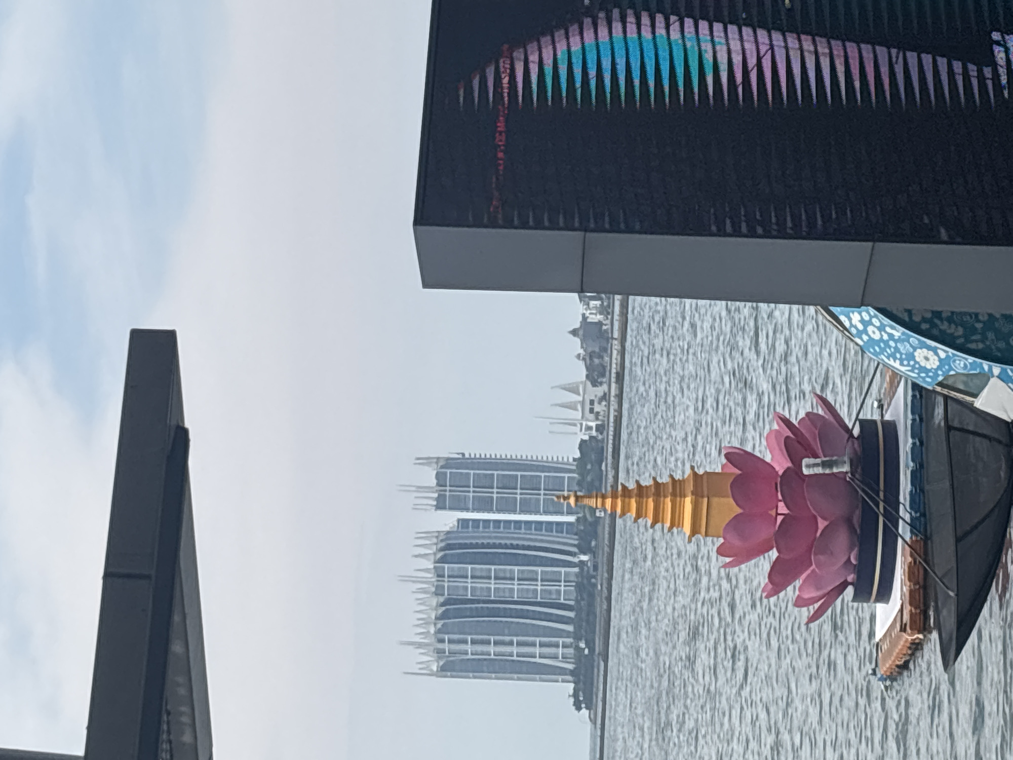 The image shows a sideways view of a scene in North Jakarta, Indonesia. The foreground features abstract architectural structures. A large digital billboard screen with colorful abstract patterns dominates the upper-right corner, and a dark geometric structure is visible on the left. In the middle ground, a body of water reflects modern high-rise buildings with curved architectural designs. A decorative float featuring a pink lotus flower and golden spire is seen on the water. The background shows a hazy sky.