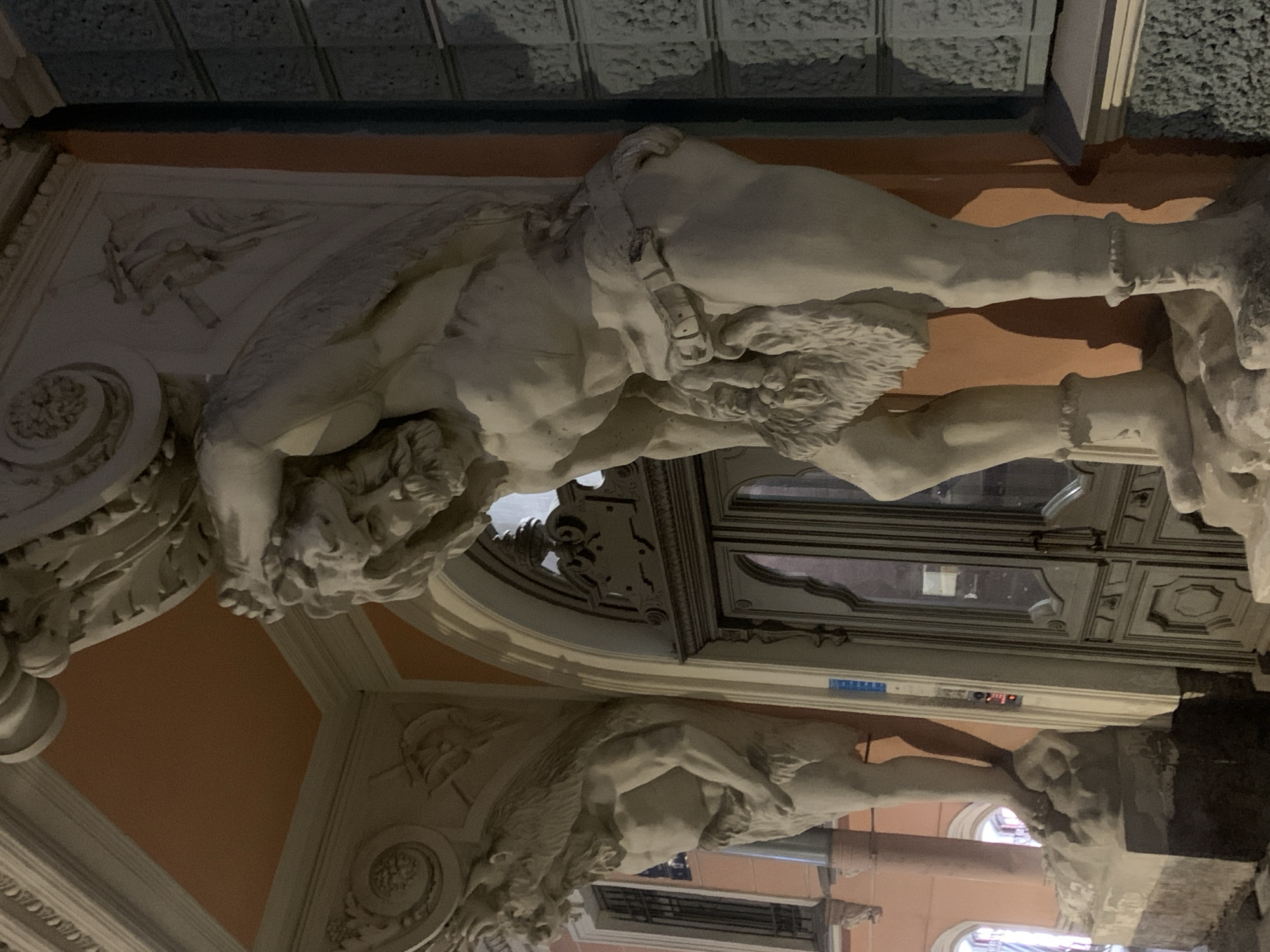 The image shows the exterior of a building at night, featuring two large stone sculptures of muscular male figures supporting an ornate architectural element above. The figure in the foreground is positioned centrally and appears to be straining under a heavy load, with his arms raised and his head bowed. He is wearing a belt around his waist and a furry loincloth. The figure on the left is partially visible and is also in a supporting pose. The building facade behind the sculptures has a large, arched wooden door with intricate carvings and metal handles. The walls are painted a warm, peachy color, contrasting with the grey stone of the sculptures and the darker, textured wall to the right. The scene suggests a classical or baroque architectural style. This is likely the Kazan Cathedral in Saint Petersburg, Russia, known for its impressive colonnades and surrounding architecture.
