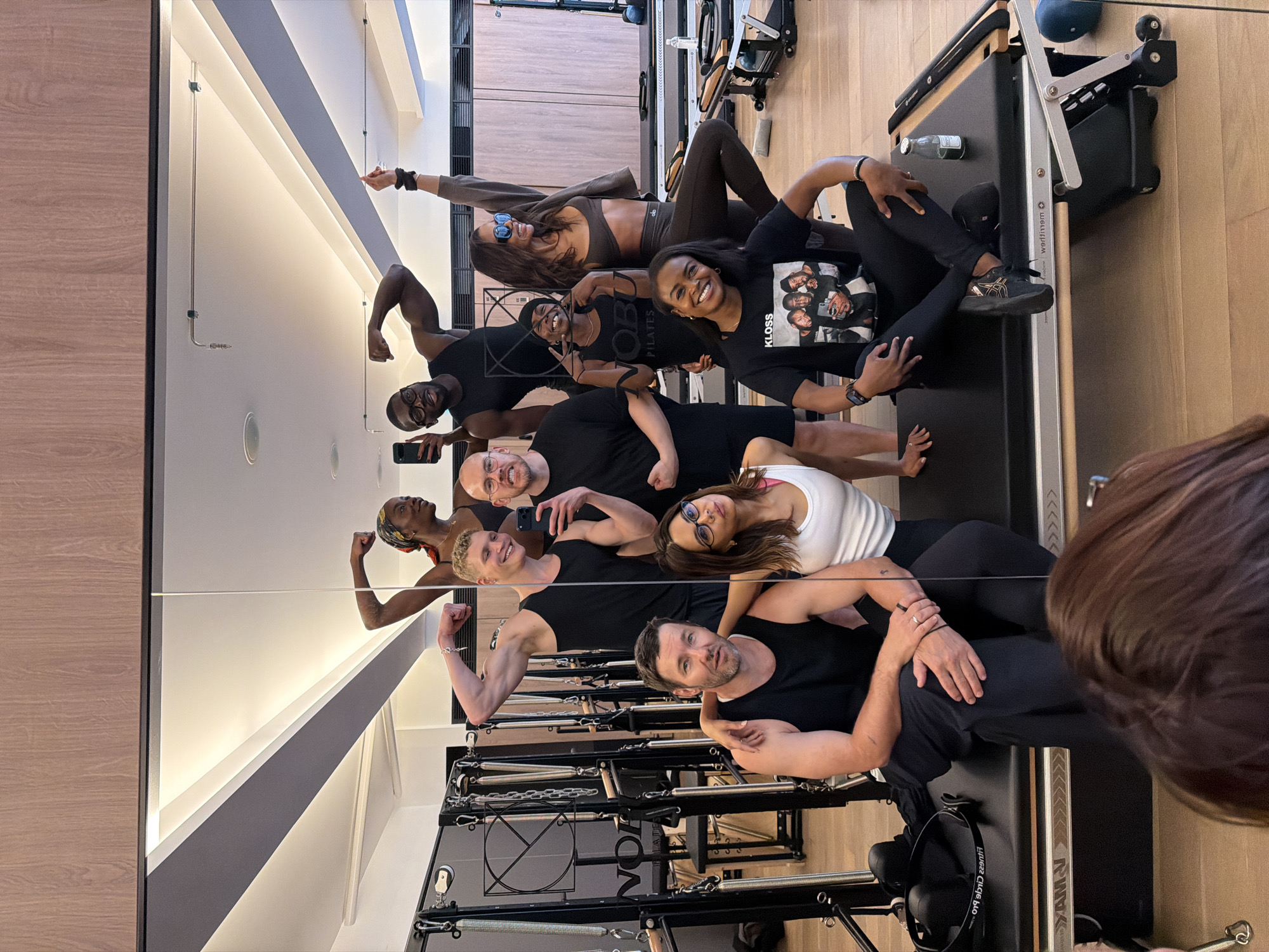 The image captures a group of nine individuals, six men and three women, posing for a mirror selfie in what appears to be a high-end pilates studio. The studio features light wood paneling, modern lighting fixtures, and specialized pilates equipment such as reformers and cadillacs. One reformer is clearly visible in the foreground, with a black mat and visible springs and handles.
The individuals are dressed in athletic attire. Several are striking poses, flexing muscles or raising arms, suggesting a lighthearted and energetic atmosphere. One person holds up a cell phone to take the photograph.
The text "NOBU PILATES" is visible on a black t-shirt worn by one of the women, and "KLOSS" is emblazoned on another t-shirt. The presence of "Nobu" suggests a connection to the luxury hotel and restaurant brand, which has locations in London. Given the upscale nature of the studio and the potential association with Nobu, the location is likely within a Nobu Hotel in London, United Kingdom, such as Nobu Hotel Shoreditch.