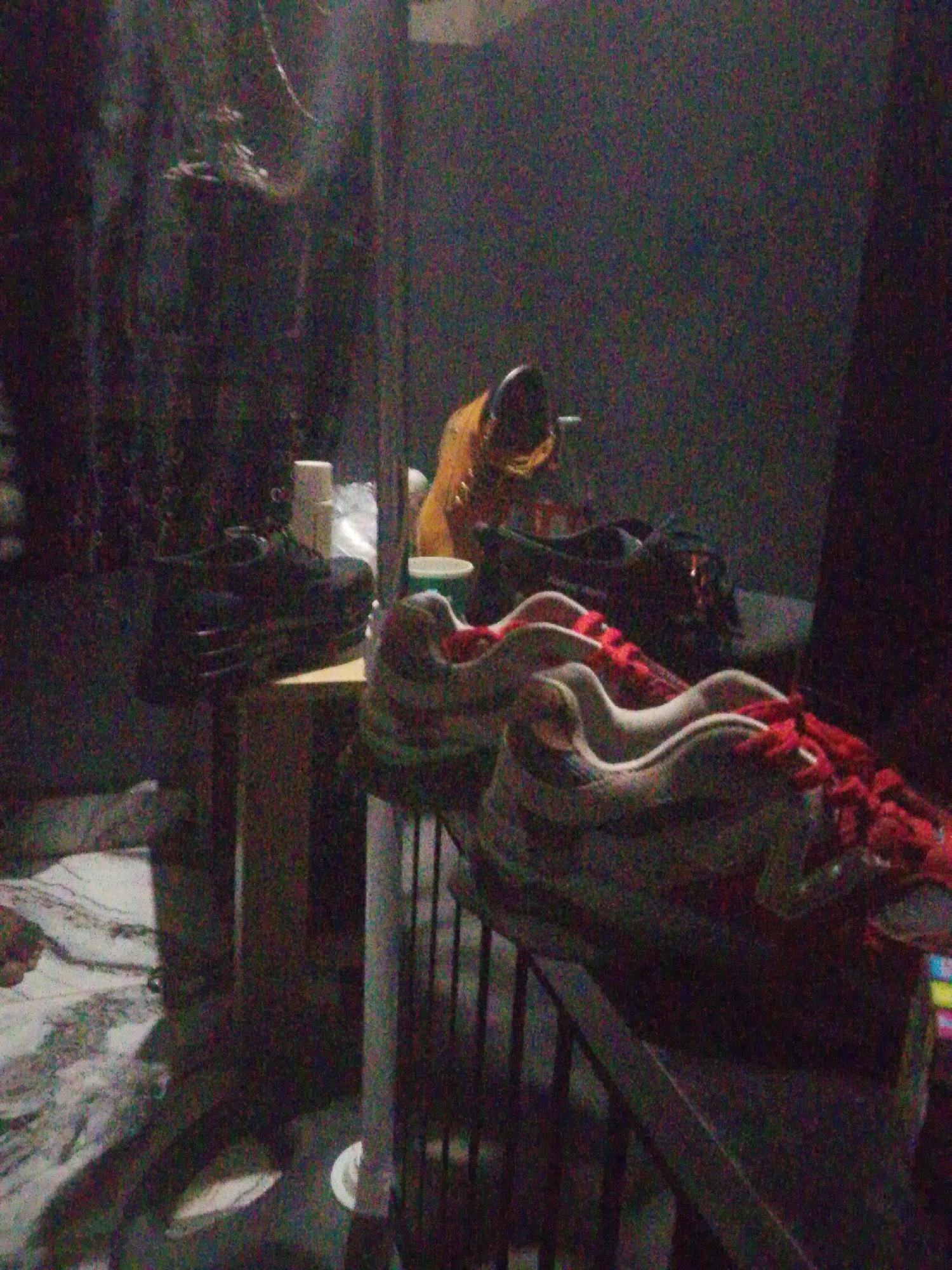 The image depicts an indoor scene, likely a room, with several pairs of shoes visible. In the foreground, a pair of white and grey athletic shoes with red laces is prominently displayed. To the left, a dark-colored shoe is placed on a wooden surface. In the background, a yellow boot and other dark shoes are present. The environment appears to be dimly lit, with some objects casting shadows. The image quality is low, with a significant amount of digital noise, making it difficult to discern finer details. The scene does not provide any specific information about the location in Keffi, Nigeria, beyond it being an indoor setting.