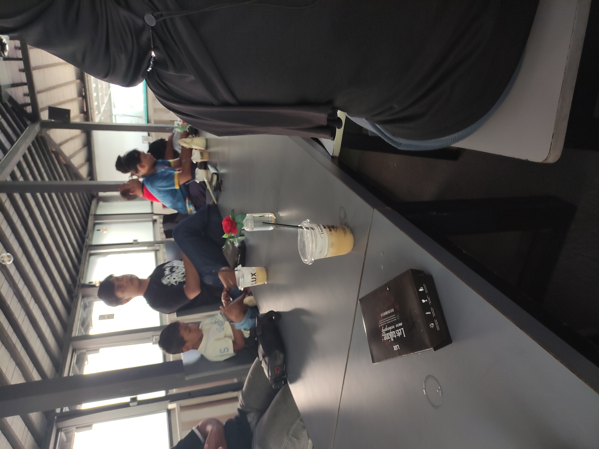The image depicts a group of people seated at a long, dark gray table in an open-air dining area with a wooden beamed ceiling. The scene appears to be at a cafe or restaurant, possibly in Kartasura, Indonesia, judging by the presence of various beverages and the attire of some individuals.
In the foreground, a black box with text and symbols is placed on the table, along with a clear plastic cup containing a light-colored beverage and a straw. To the left of this, a black bag is situated on the table. Further down the table, another clear plastic cup with a beverage and straw is visible, alongside a small vase containing a red flower and water.
Several individuals are present. On the left, a person in a black shirt and gray pants is partially visible. Next to them, a young boy in a light-colored shirt is seated. Further down, a man in a black t-shirt is seated. In the background, a person in a blue shirt with yellow trim is visible, as is another individual wearing a black headscarf. The lighting suggests it is daytime, with natural light filtering in from the sides. The overall atmosphere seems relaxed, with people engaged in conversation or enjoying their drinks.