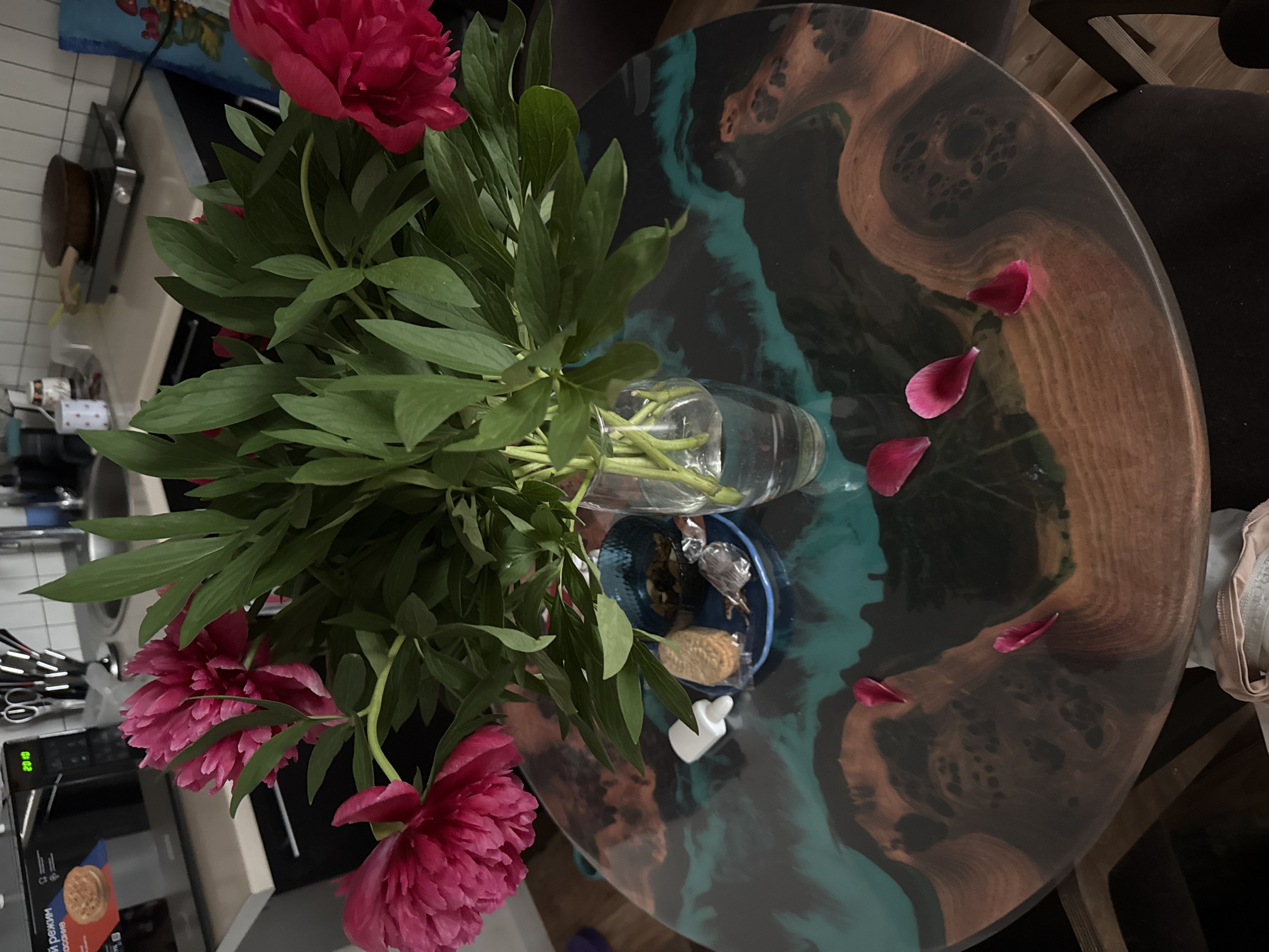 The image shows a bouquet of dark pink peonies in a clear glass vase placed on a round table with a unique, patterned surface that appears to be resin art. The table has swirls of brown and teal hues. Several fallen peony petals are scattered on the table's surface. Beside the vase is a small blue dish containing various items. The surrounding environment includes a kitchen counter with utensils and a microwave, a dark chair, and what looks like a portion of a painting. The location within Perm, Russia is not identifiable from the image.