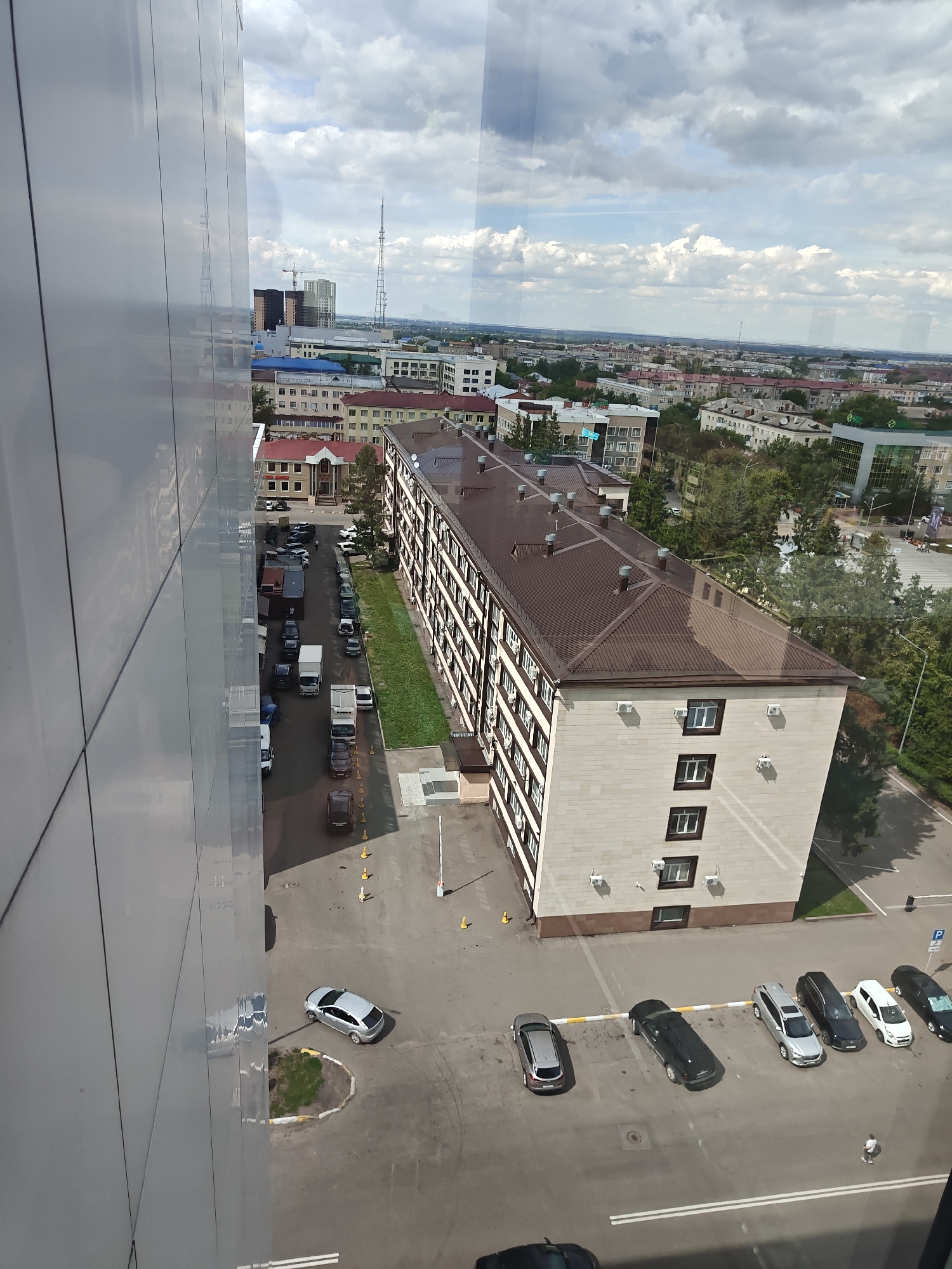 The image, taken from an elevated vantage point, depicts an urban scene in Petropavl, Kazakhstan. In the foreground, the reflective facade of a modern building dominates the left side. To the right, a long, multi-story building with a brown pitched roof and beige stone exterior is visible, with numerous windows and air conditioning units. A parking lot runs alongside this building, filled with various cars including sedans and SUVs, as well as several white trucks parked in a row. The street is lined with trees, and further into the background, a cityscape unfolds with more buildings, some taller than others, a radio tower, and a horizon under a cloudy sky. A few individuals are seen walking in the scene, one near the parked cars and another in the distance. The image appears to be taken during daylight hours.