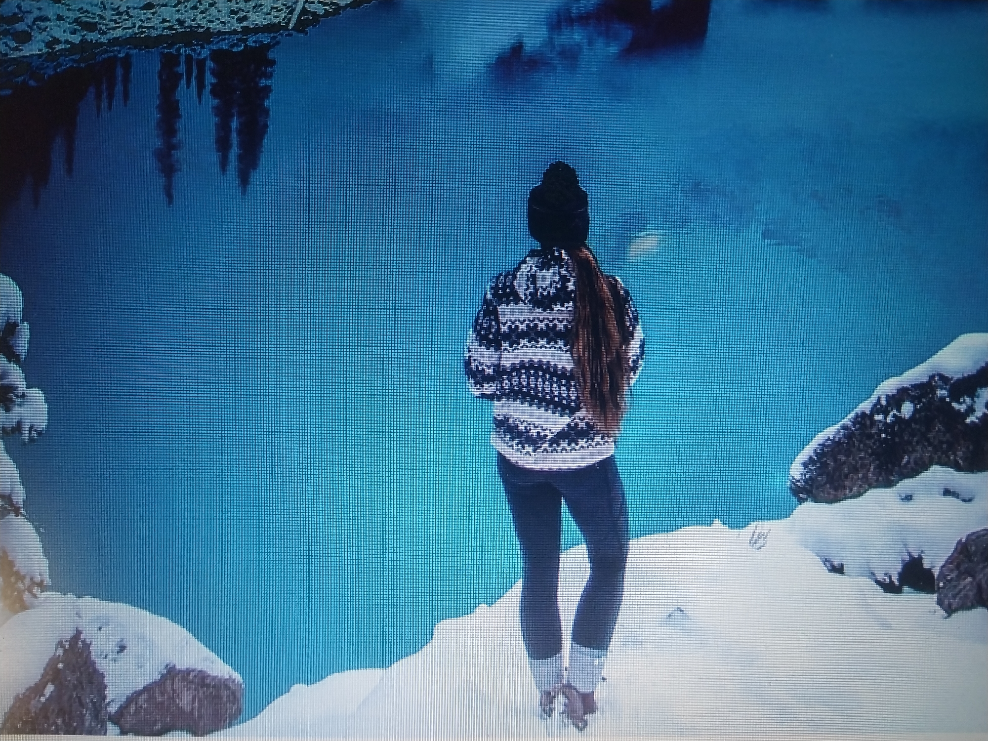 The image depicts a person from behind, wearing a black beanie, a patterned sweater, and dark pants, standing on a snow-covered landscape. The person's long brown hair is visible. The foreground shows snow-covered rocks and ground. In the background, a body of water with a deep blue hue reflects the silhouette of trees, suggesting a natural, likely cold environment, possibly a lake or a calm sea. The sky is not visible, but the overall impression is of a winter scene.